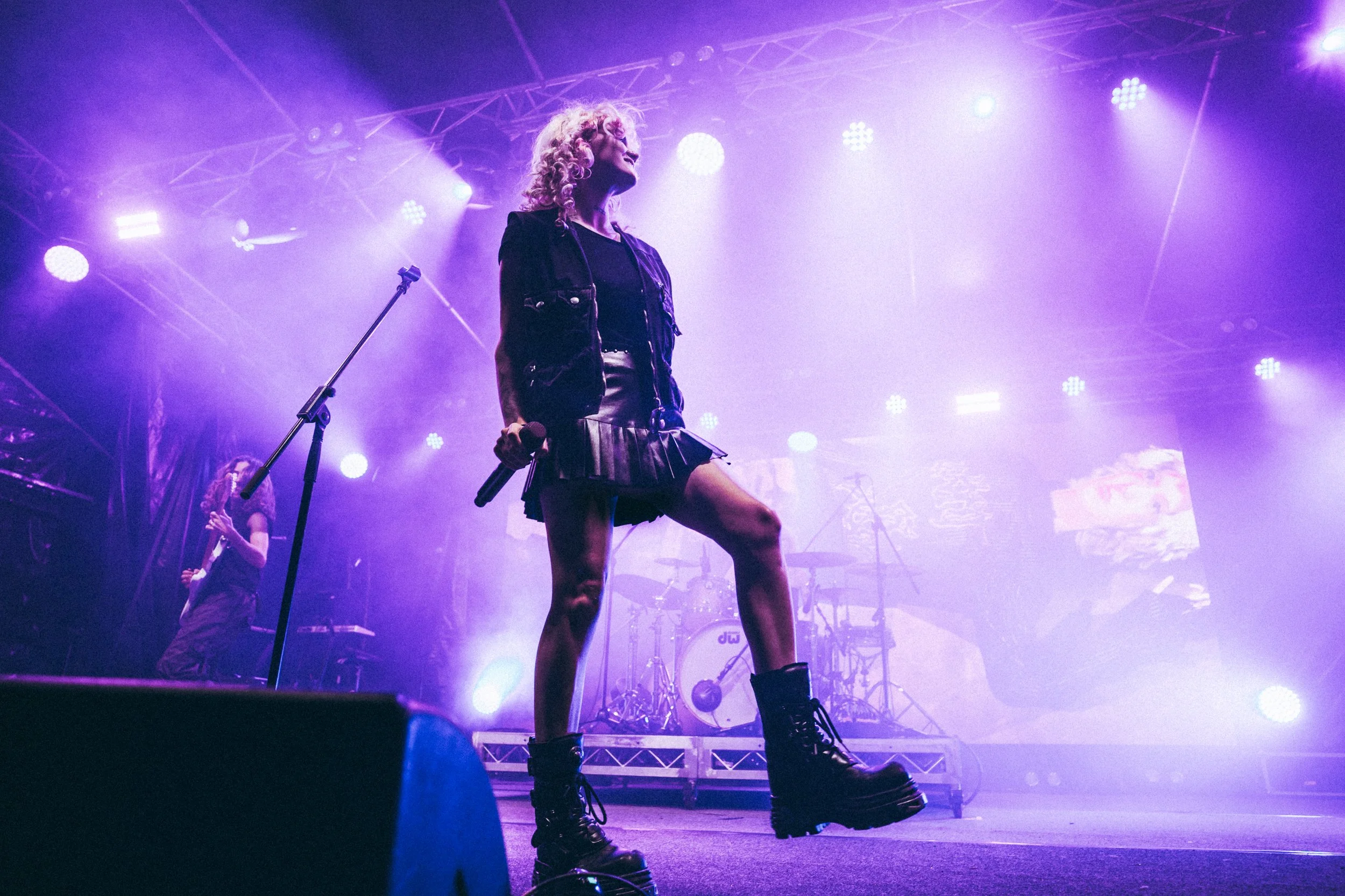 A performer with curly blonde hair wearing a black jacket and skirt, holding a microphone, stands on stage under purple and white stage lights, with another performer in the background and musical equipment around.
