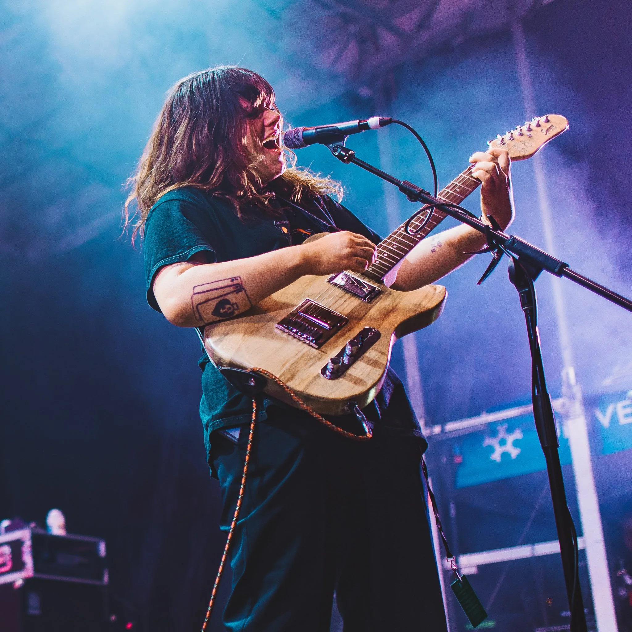 A woman with long hair and sunglasses singing into a microphone while playing an electric guitar on stage amidst colorful lights.