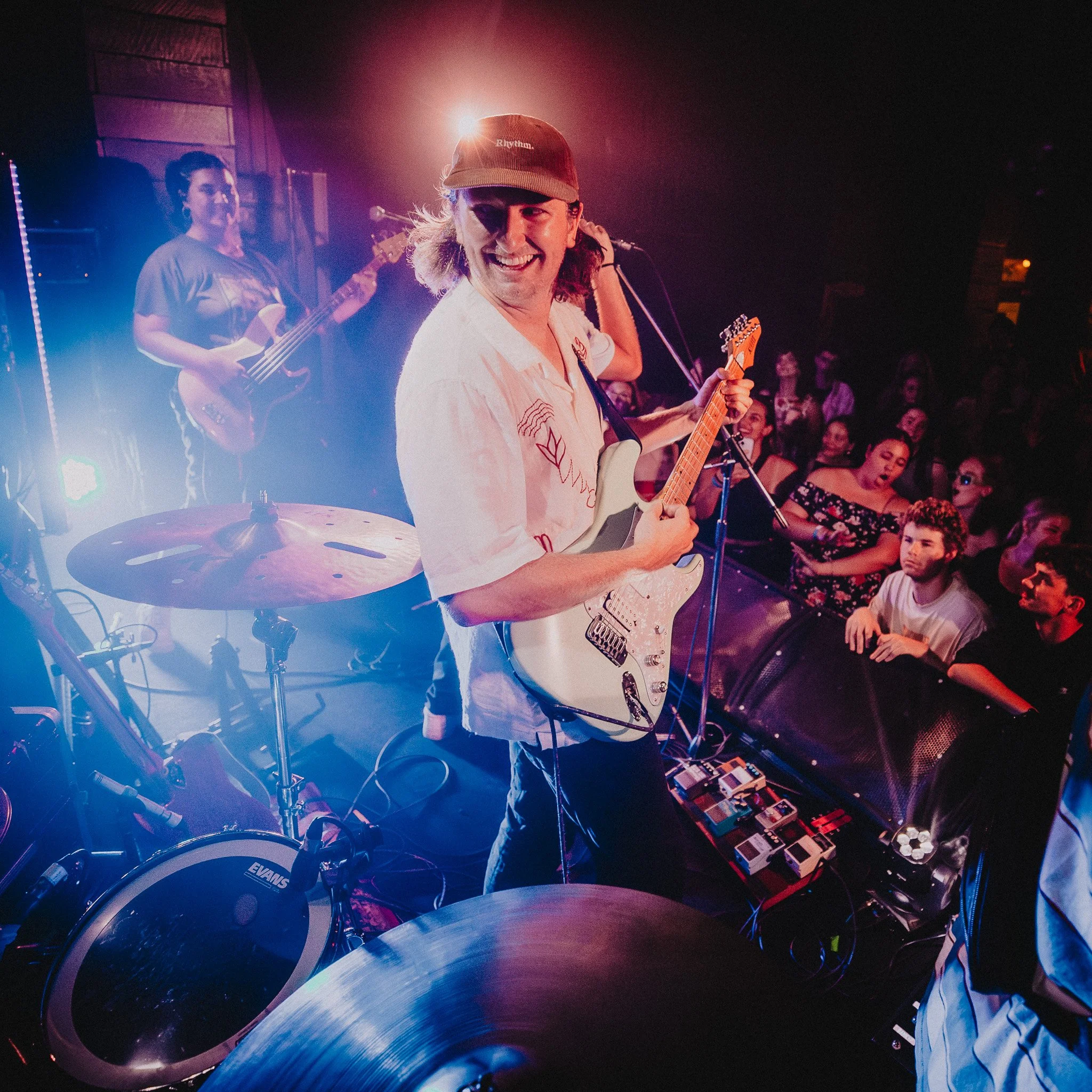 A musician with long hair wearing a cap and white shirt playing an electric guitar on stage with a smiling woman and audience in the background, colorful stage lighting.