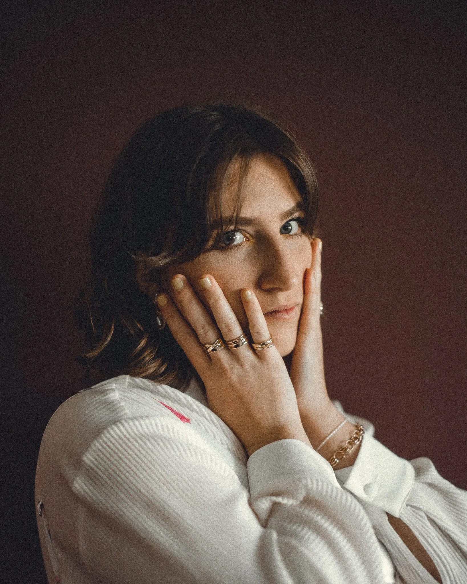 A woman with dark brown hair and blue eyes resting her face on her hands, wearing rings and bracelets, with a soft neutral background.