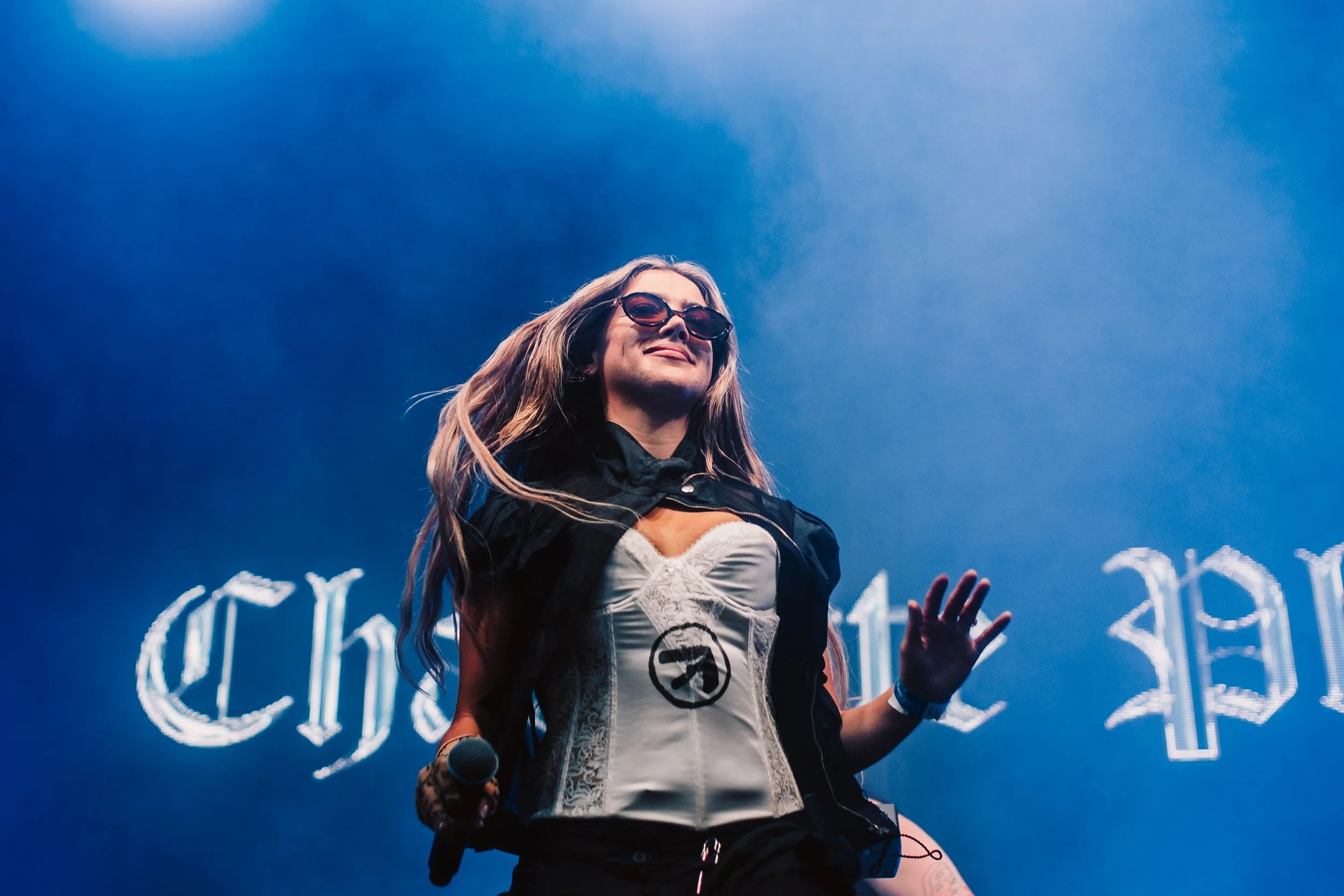 A woman with long hair wearing sunglasses and a black jacket, holding a microphone, standing in front of a blue neon-lit sign.