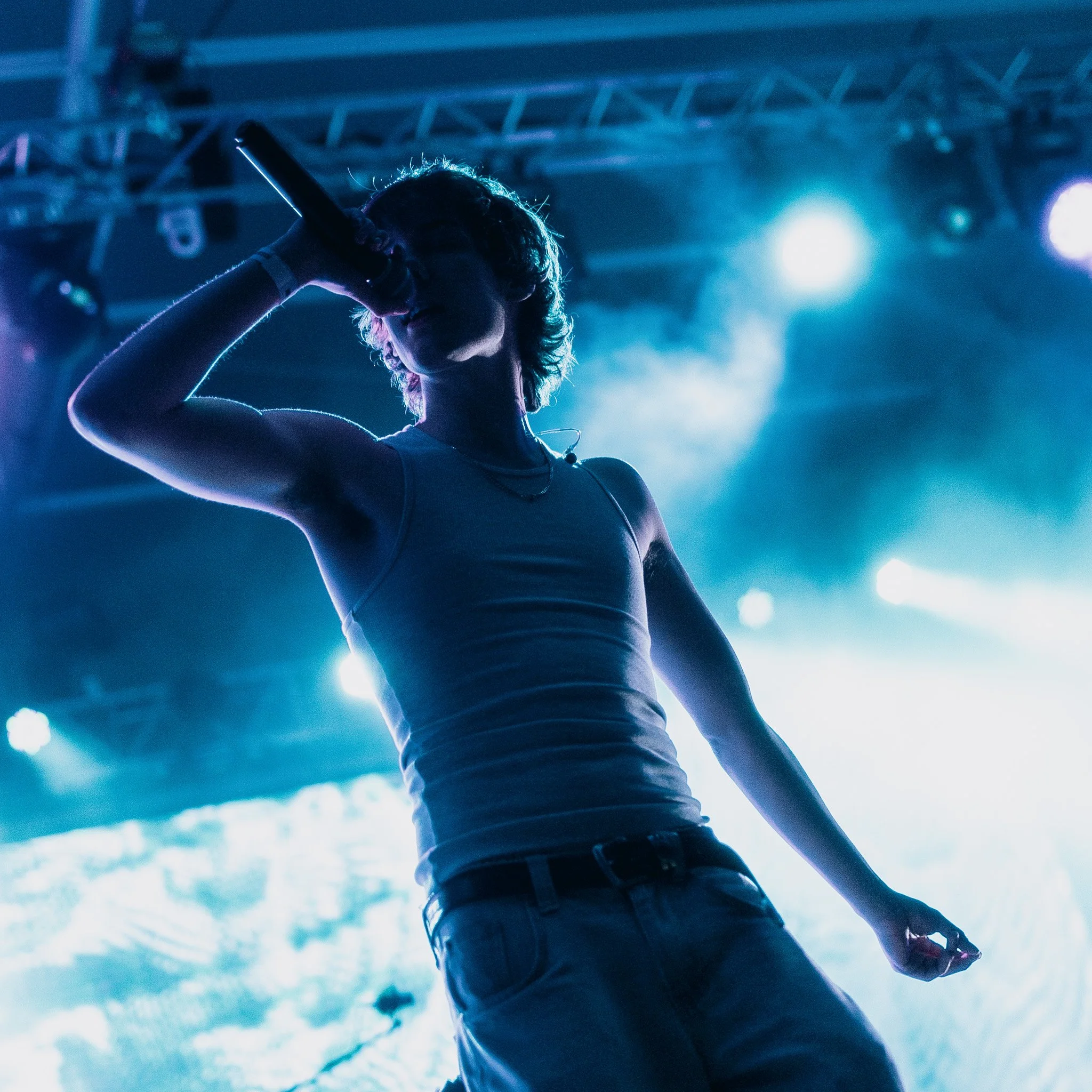 A person holding a microphone on stage with blue and purple lighting, wearing a tank top and jeans, in a concert or performance setting.