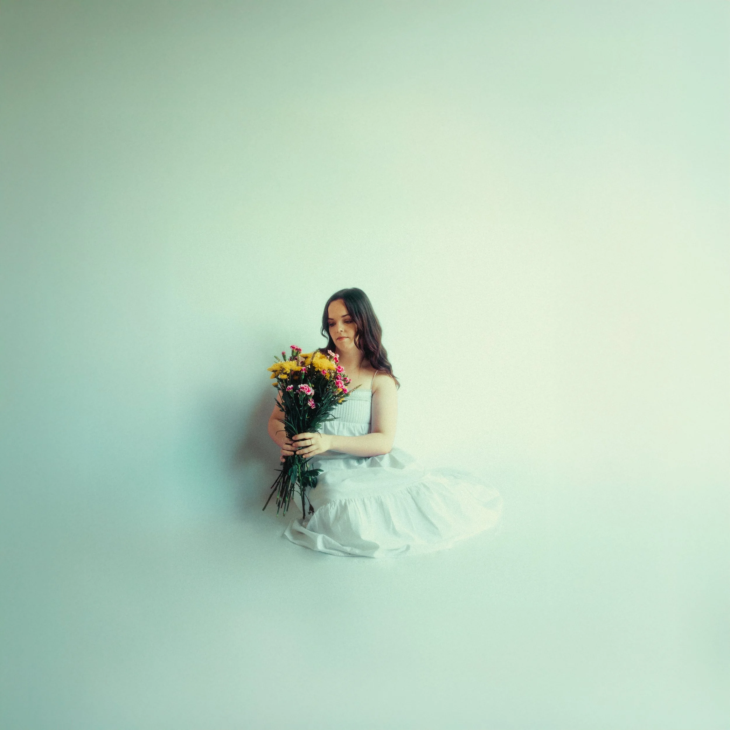 A young woman with long dark hair and light skin, wearing a white sleeveless top and a long white skirt, is sitting against a pastel green wall. She is holding a large bouquet of yellow, pink, and purple flowers and looking down at them.