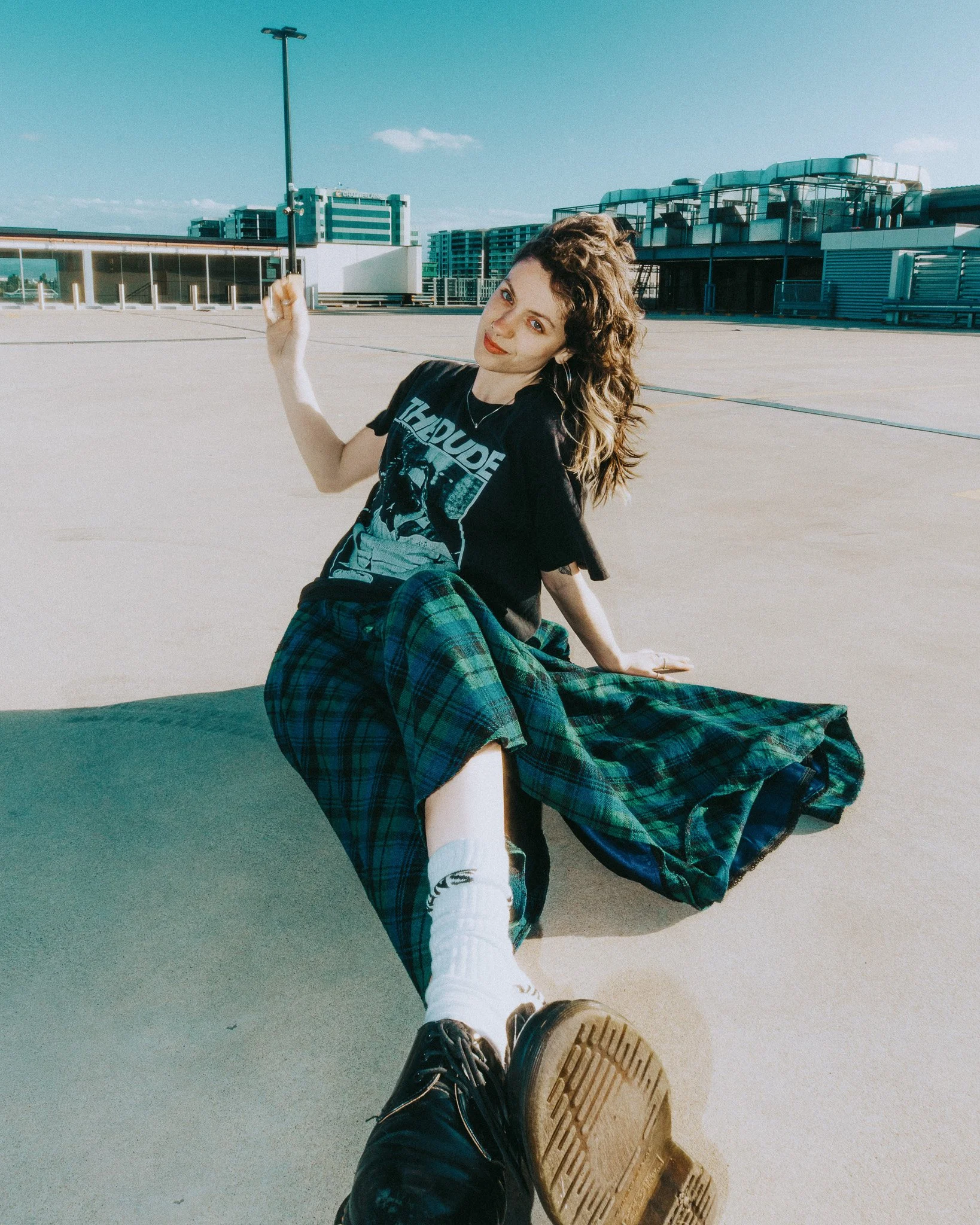 Person with curly hair sitting on a rooftop, holding up a finger, with city buildings in the background.
