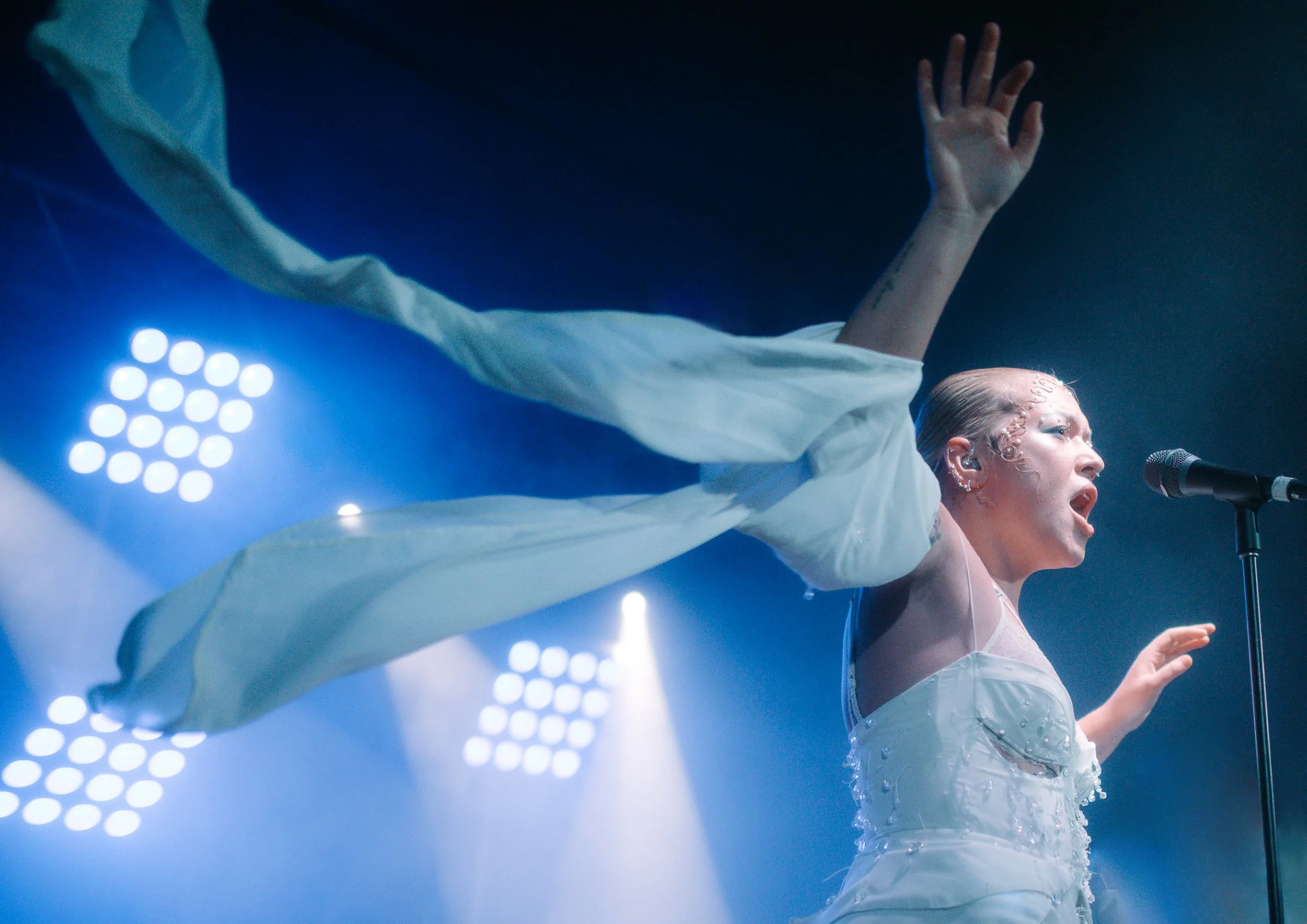 A performer on stage in a white outfit with water splashing on her from below, raising her arm and singing into a microphone, with stage lights illuminating the scene.