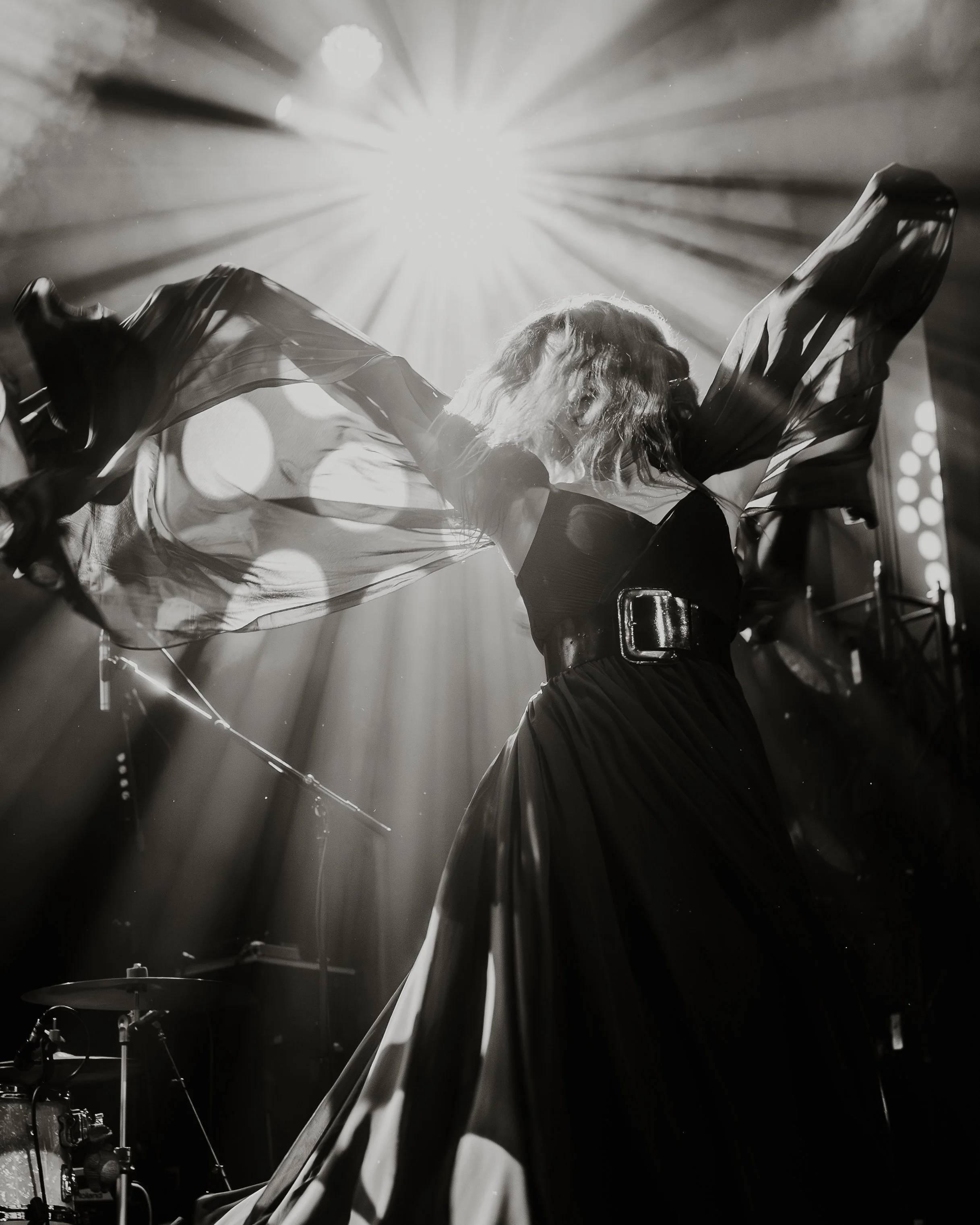 A musician performing on stage with dramatic lighting, backlit by a bright light source creating rays and silhouettes, with musical instruments visible in the foreground.
