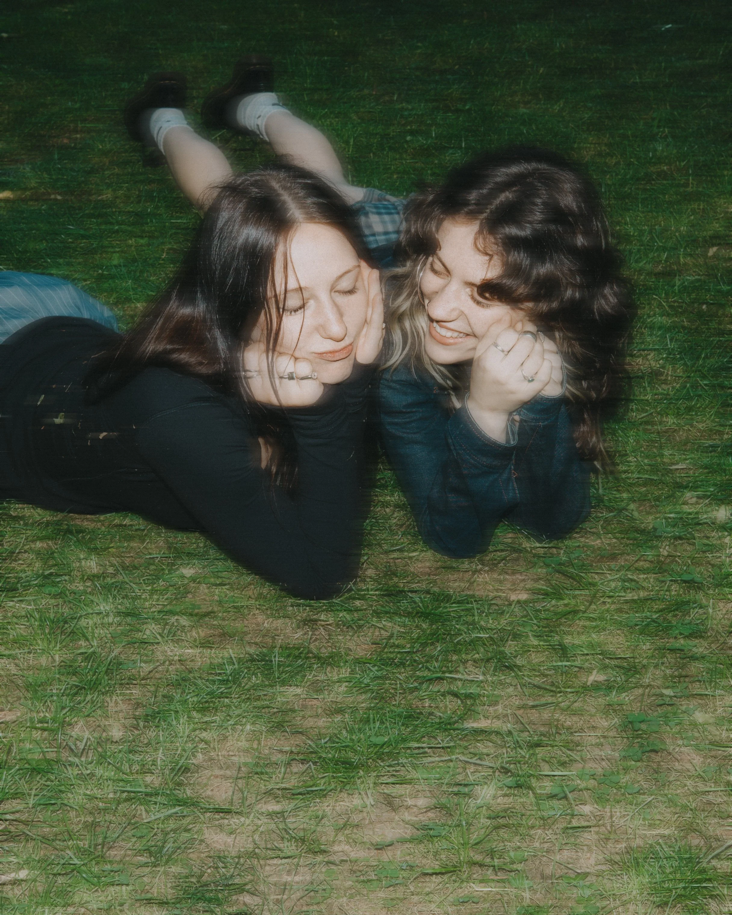 Two women lying on green grass, facing each other and smiling, with their heads close together and hands resting on their cheeks. One has long dark hair, and the other has wavy dark hair.
