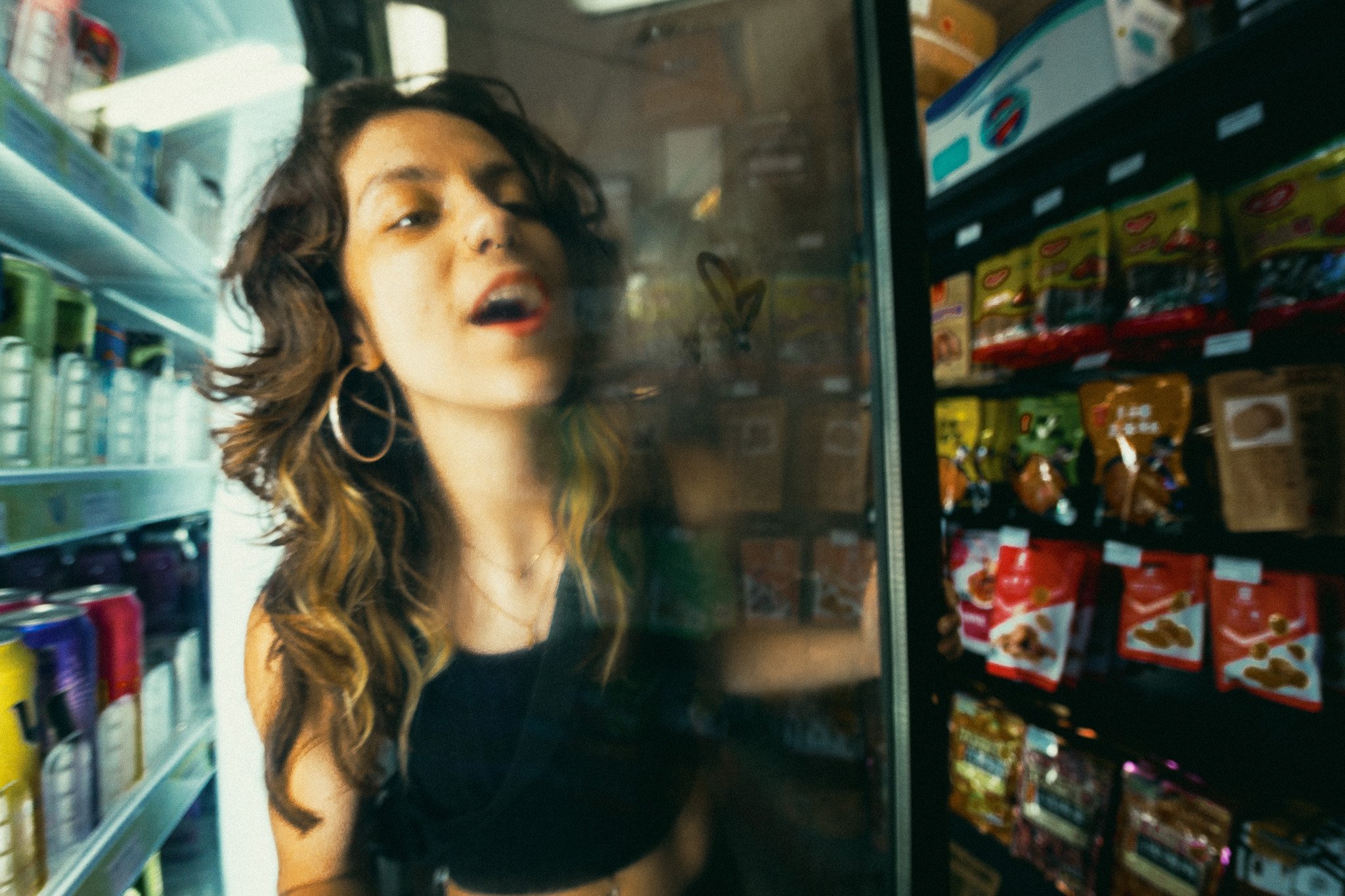 A young woman with dark curly hair and hoop earrings looking through a supermarket refrigerator door.