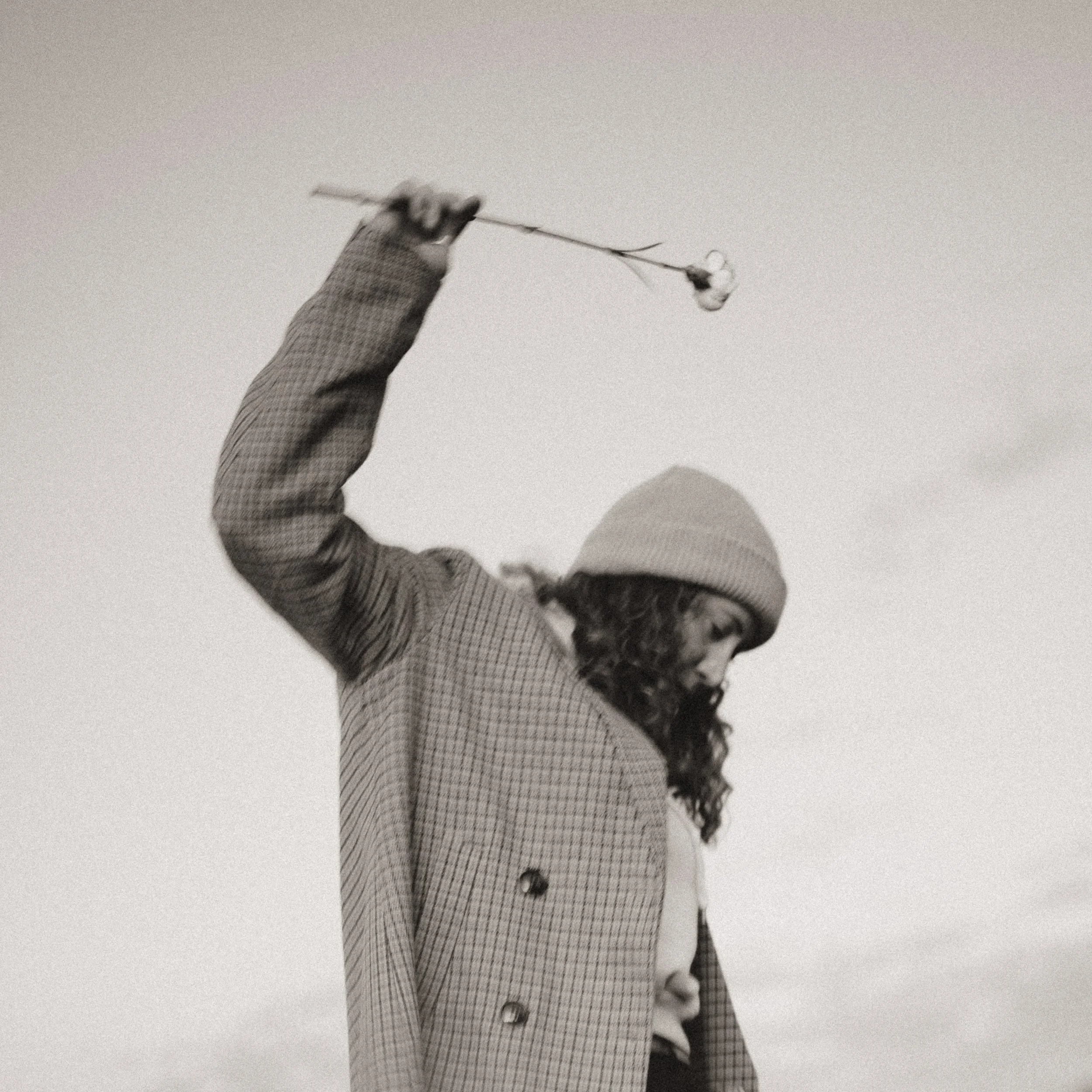 Black and white photo of woman with long hair, wearing a checkered blazer, holding a long stick with a cotton flower at the end, against a plain background.