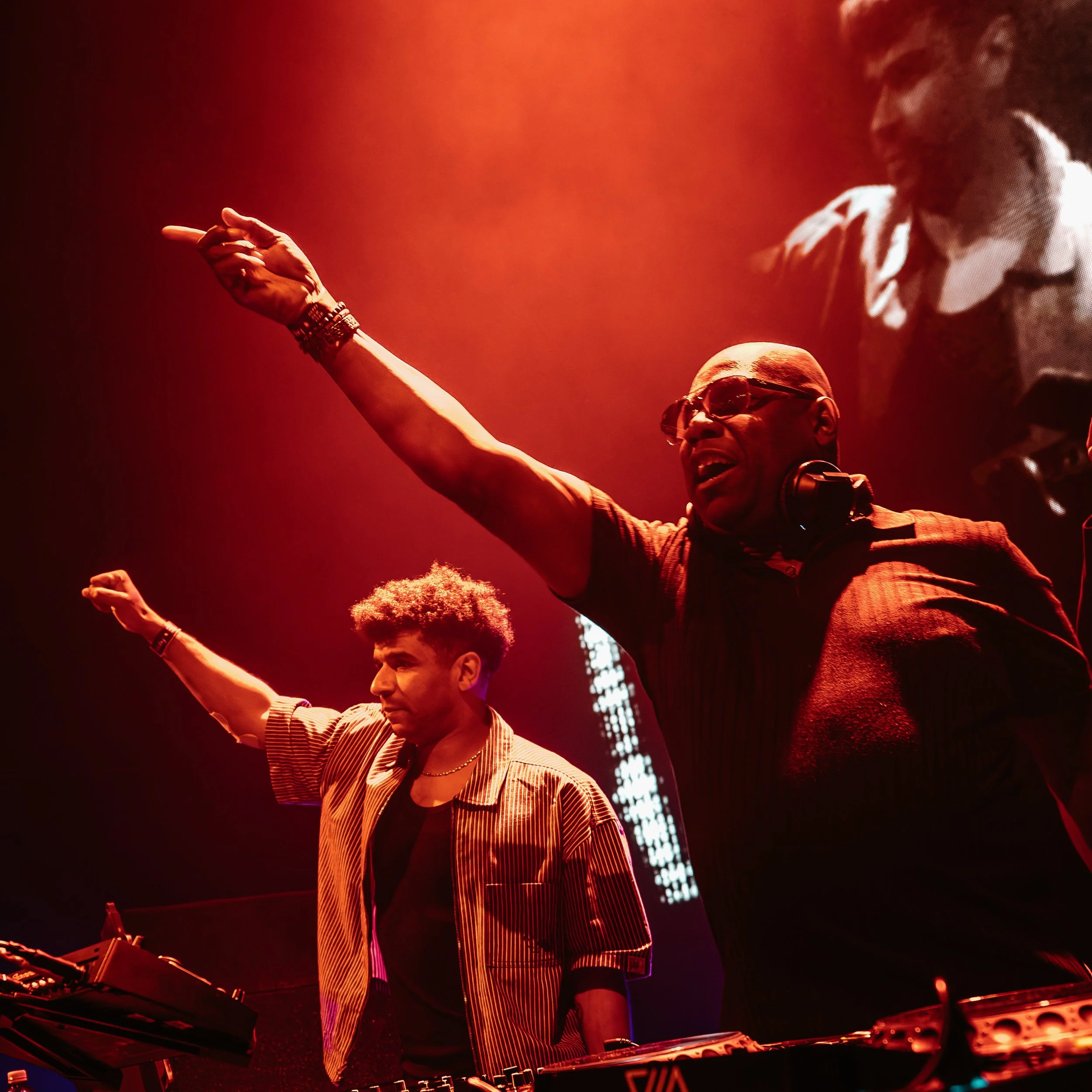DJ performing on stage with red lighting, wearing sunglasses and striped shirt, surrounded by musical equipment.