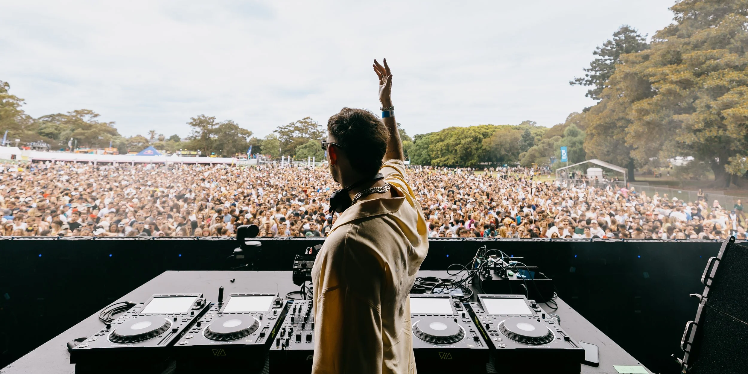 DJ in beige jacket performing at outdoor music festival with large crowd of people and trees in the background.