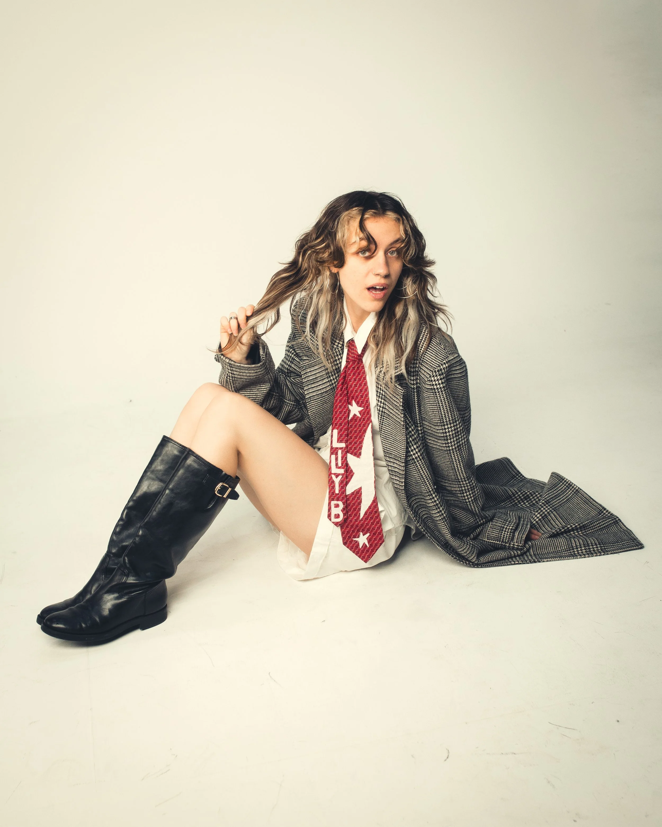 A young woman with curly hair and dressed in a plaid blazer, tie, and white shirt, sitting on the floor, one leg up, wearing black knee-high boots, against a plain white wall.