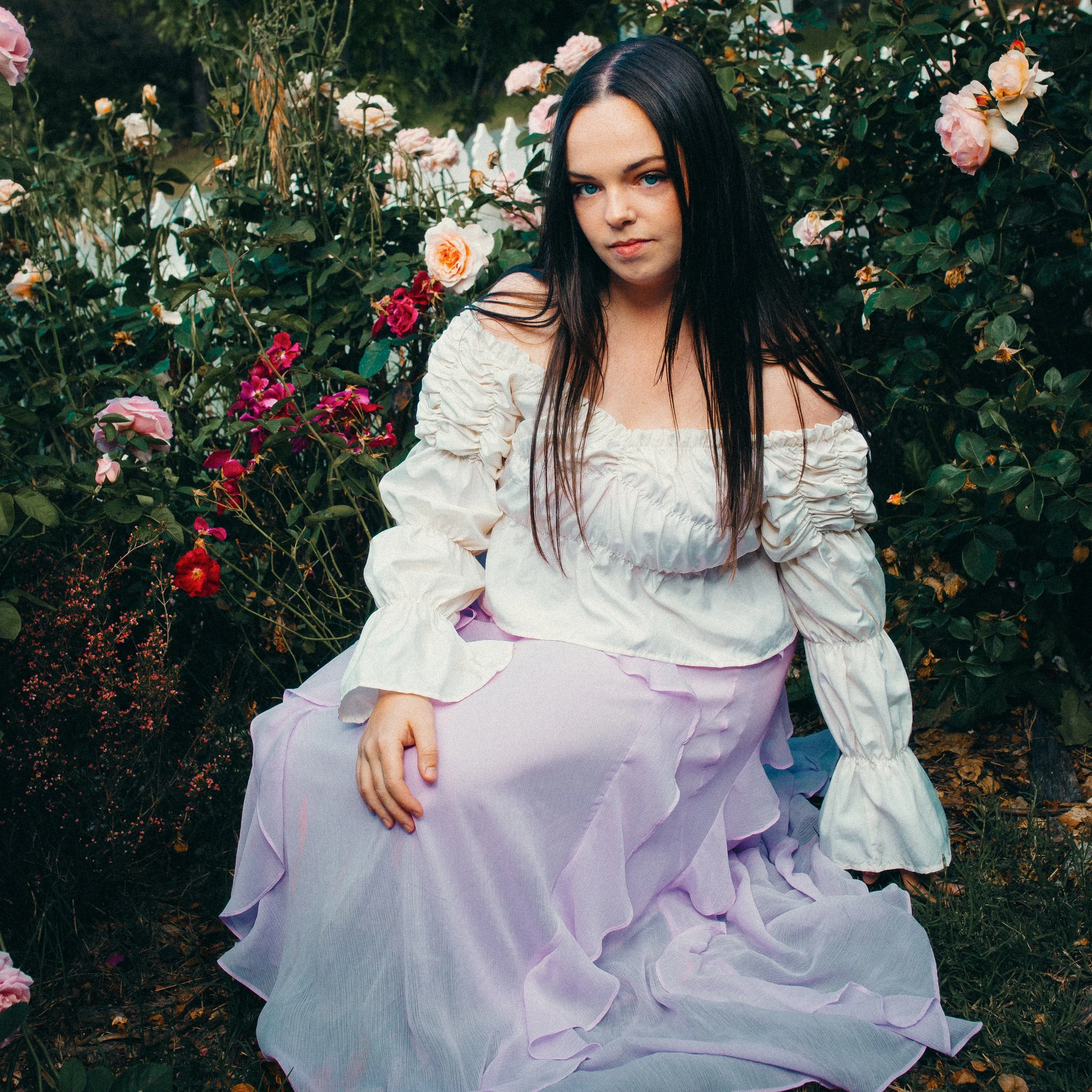 A young woman with long dark hair and blue eyes standing among pink and white roses in a garden, wearing a white off-the-shoulder dress.