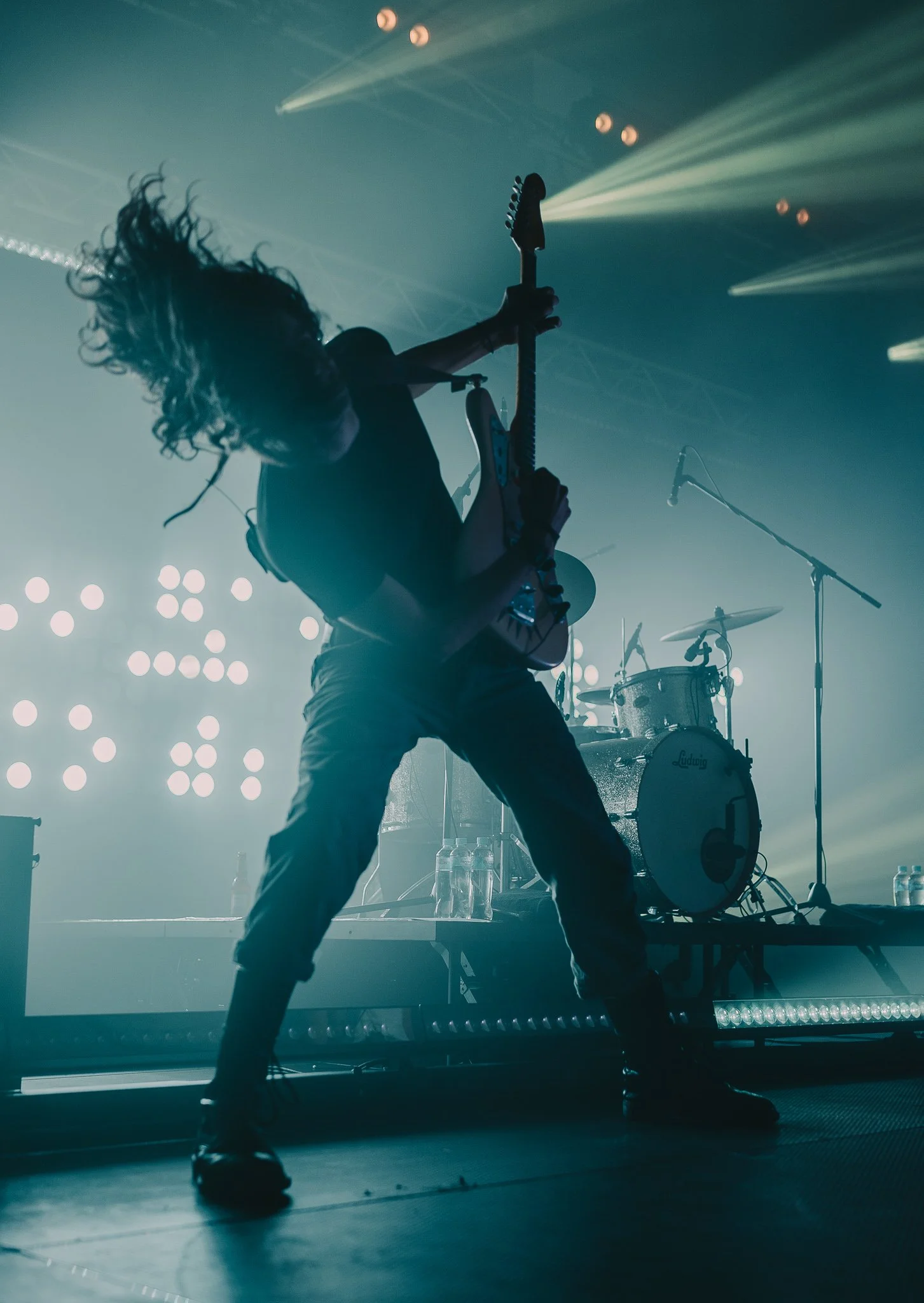 Silhouette of a guitarist performing on stage with dramatic lighting, drums in the background, and stage equipment.