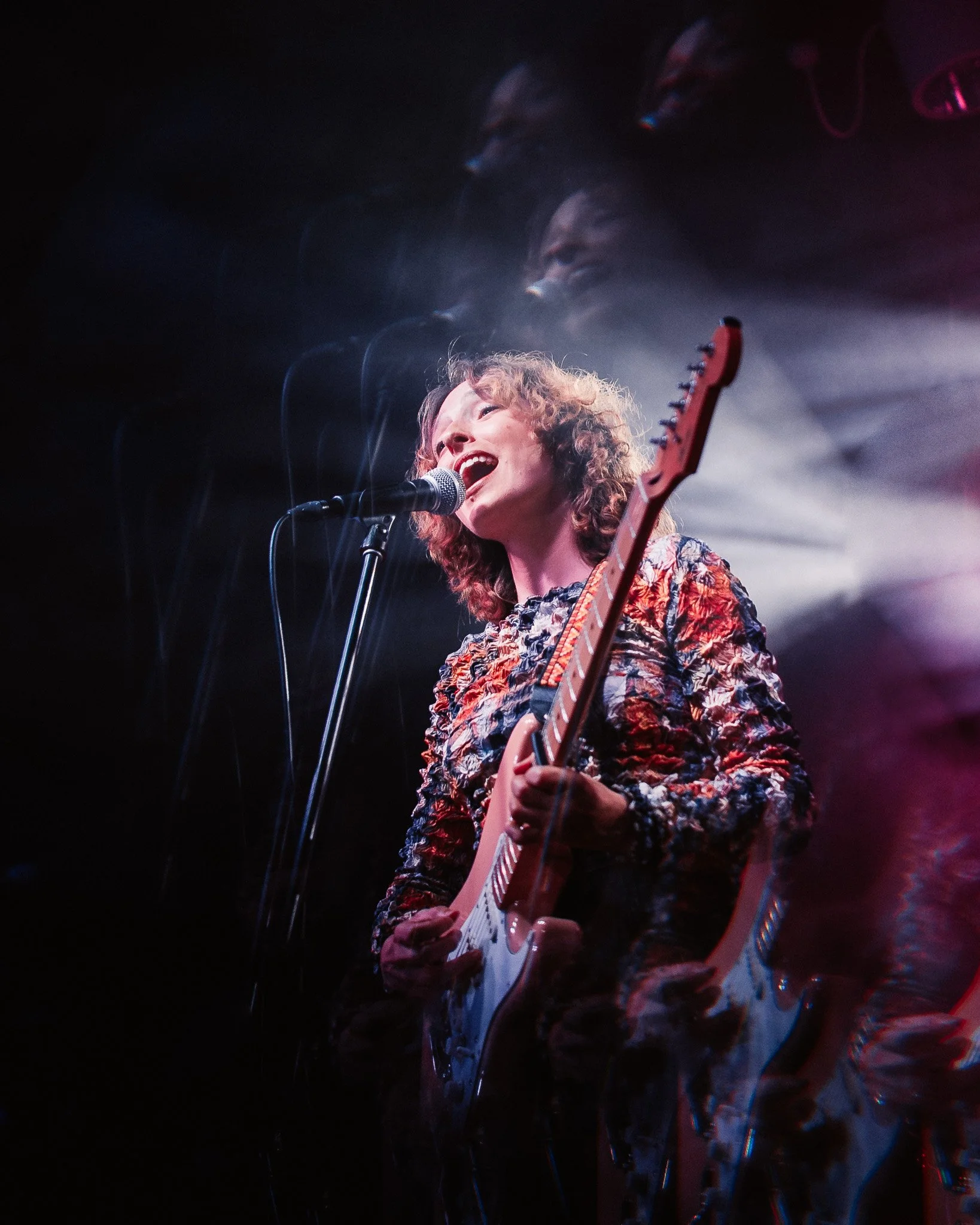 Female singer with curly hair performing passionately on stage, playing an acoustic guitar and singing into a microphone with colorful stage lighting creating a blurred, abstract background.