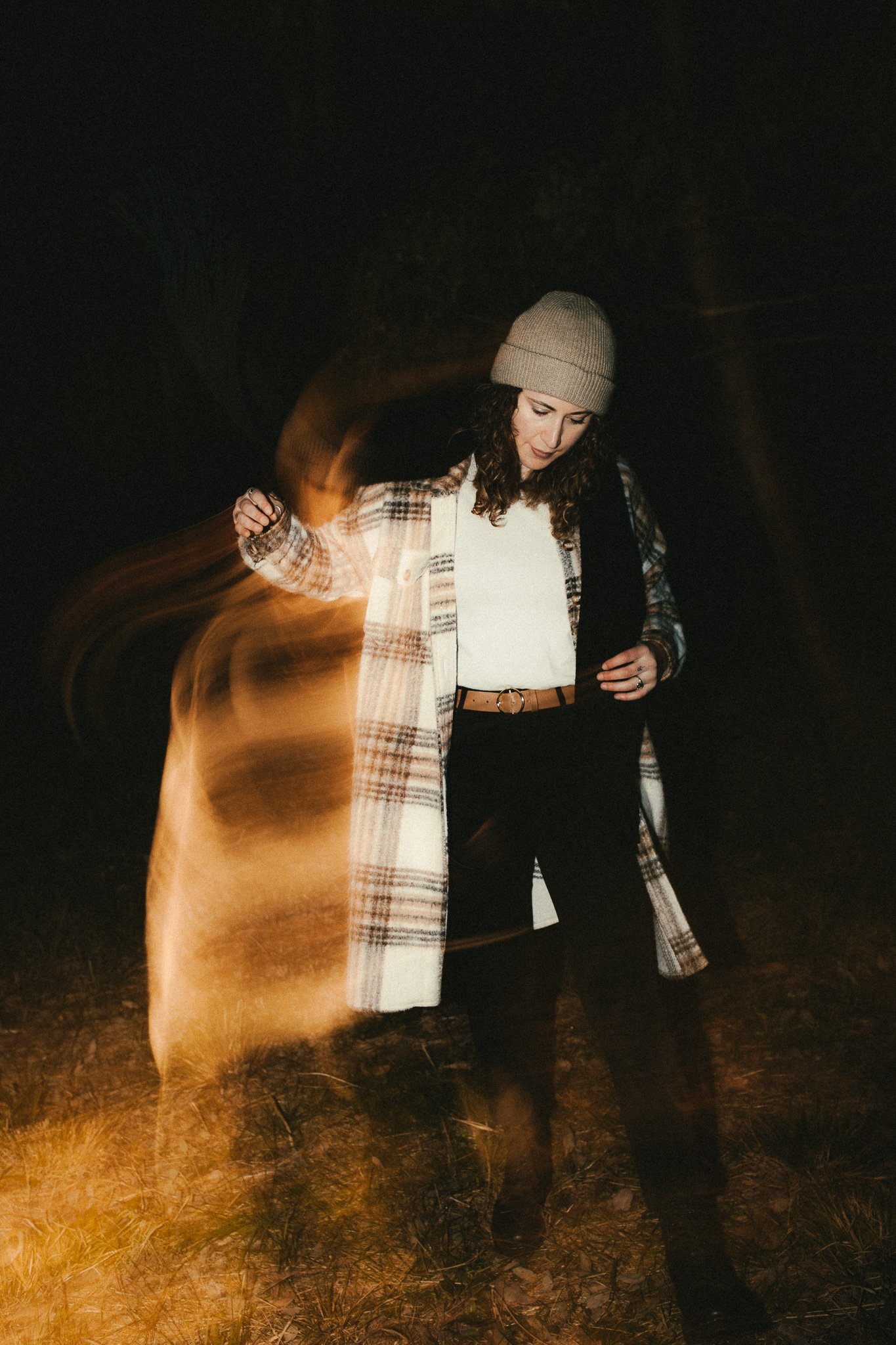 A woman with curly hair wearing a beige knit beanie, a plaid coat, white top, black pants, and black boots stands in a dark outdoor setting at night. She appears to be looking down with her head slightly tilted.