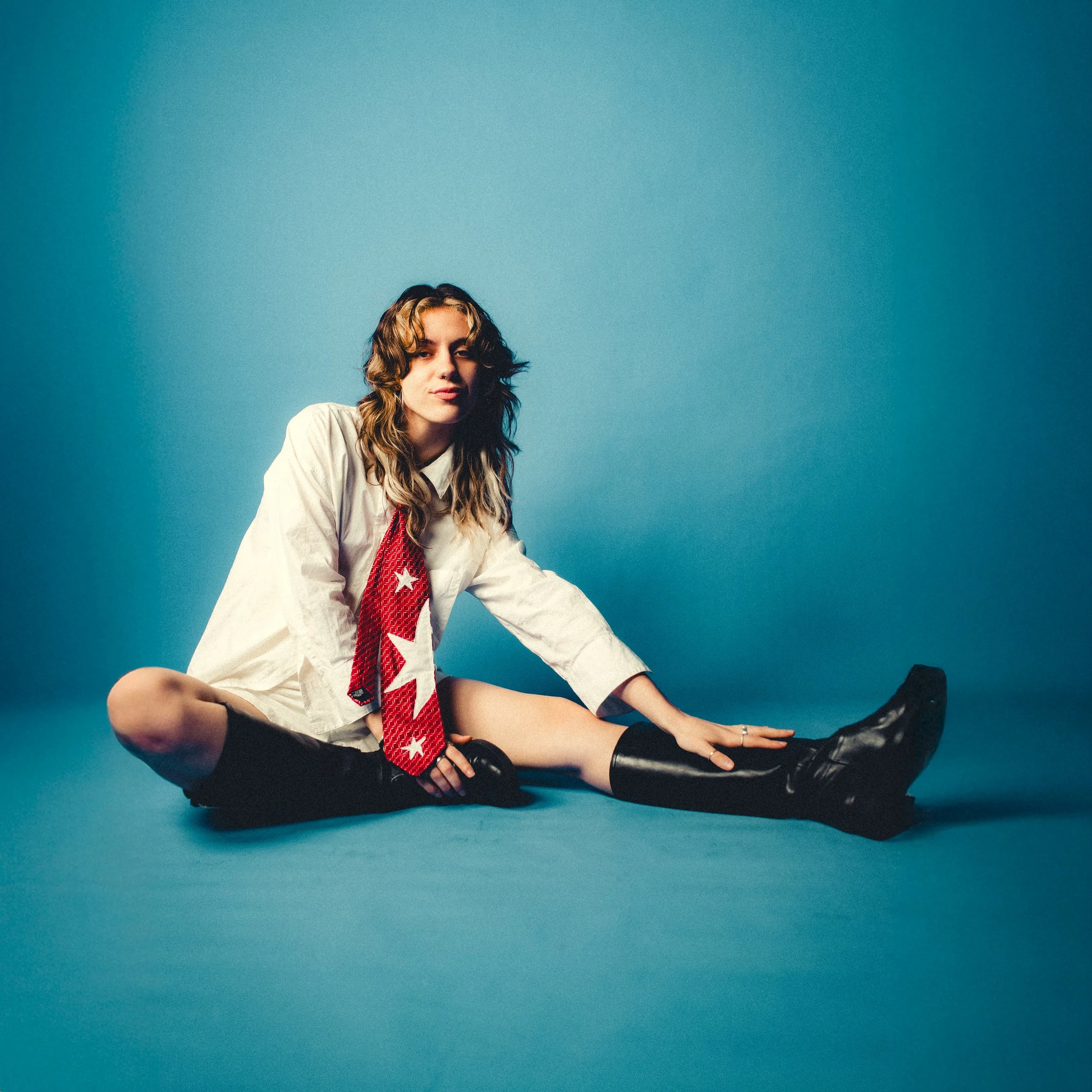 A young woman with wavy brown hair sitting on the floor against a blue background, wearing a white shirt, a red and white star-patterned tie, black shorts, and black boots.