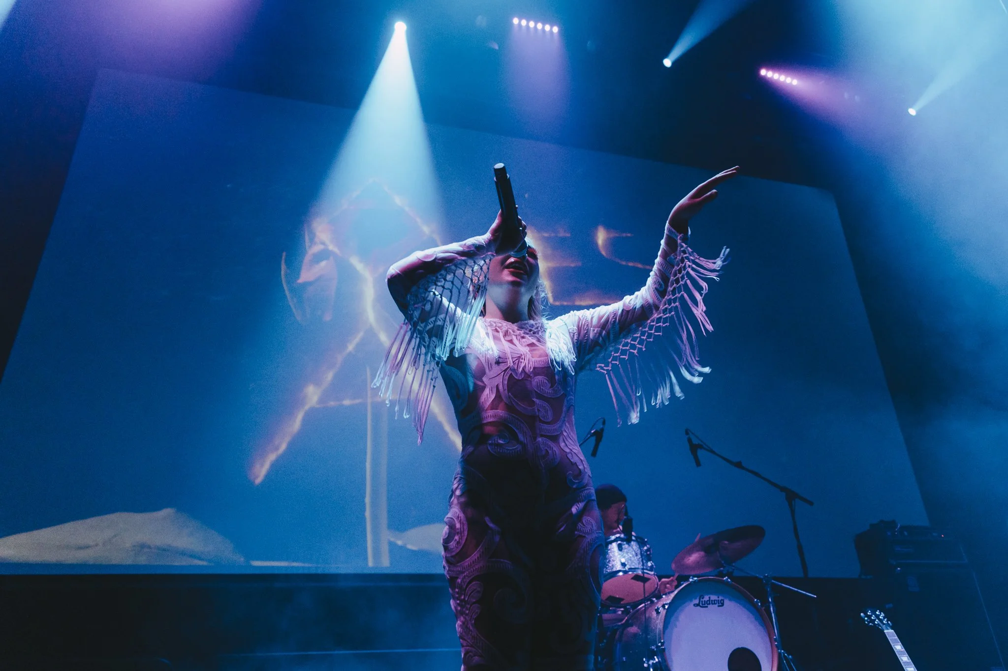 A singer with long hair and fringed clothing performing on stage, illuminated by blue and purple stage lights, with band instruments visible in the background.
