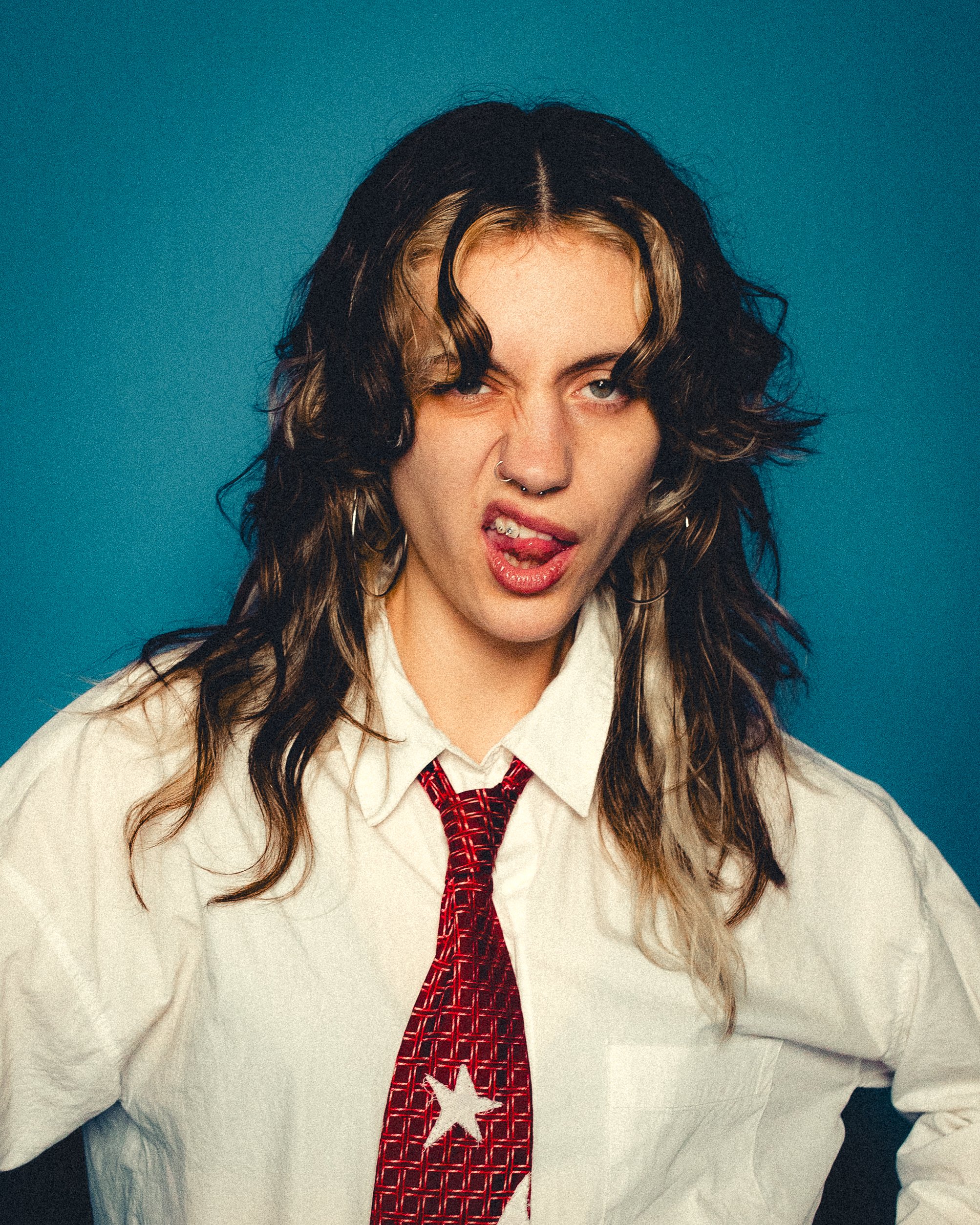 Close-up of a young woman with black and blonde curly hair wearing a white shirt and red patterned tie with a white star, making a playful facial expression with tongue out and one eye closed.
