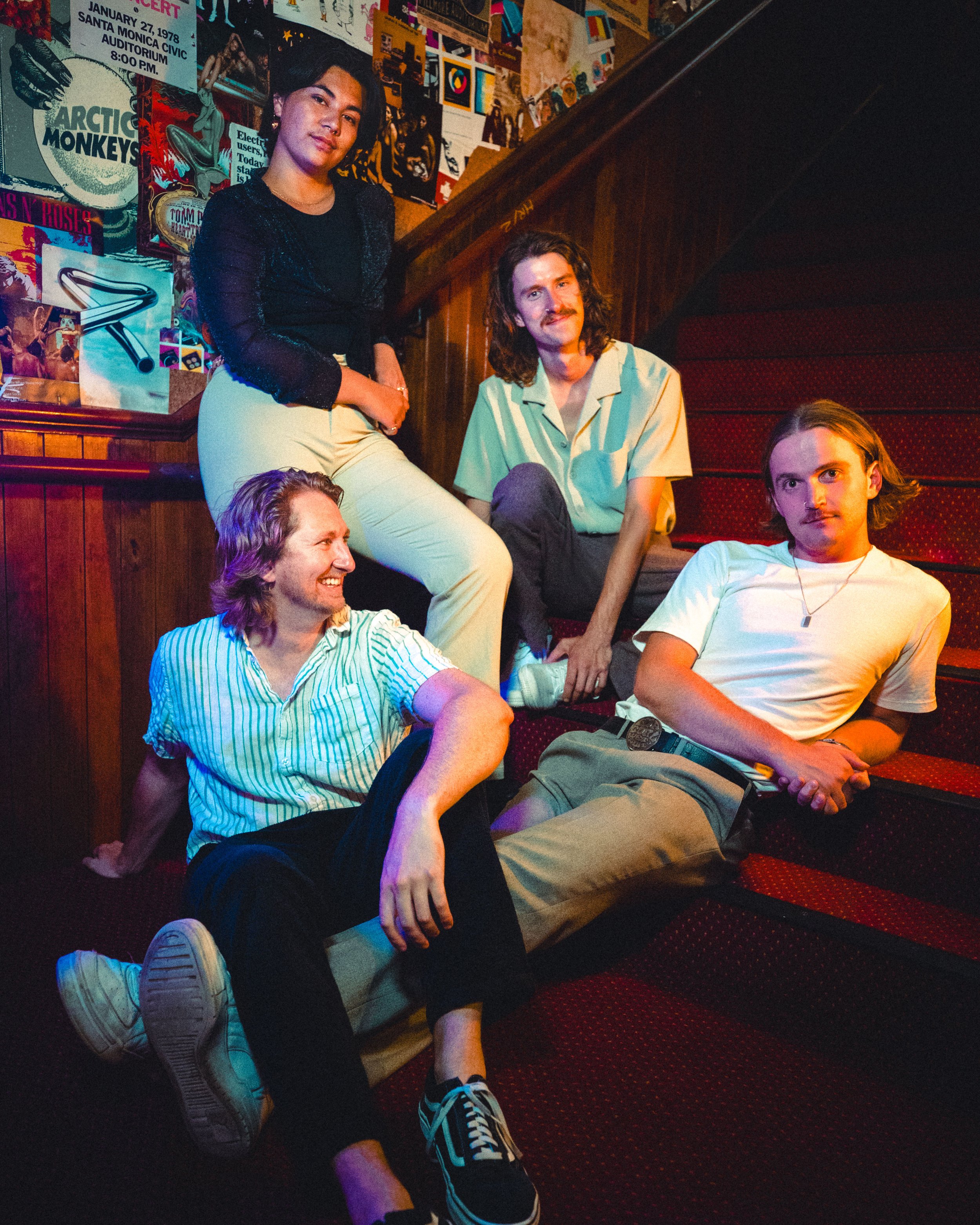 Group of five young adults sitting on a staircase inside a room decorated with posters and artwork on the walls. Four are sitting on the stairs and one is standing.