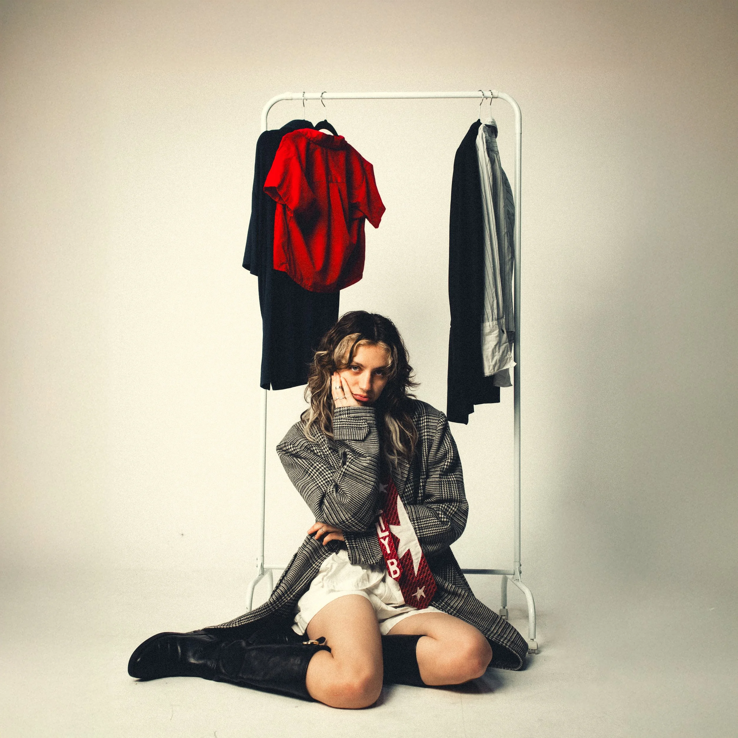 A young woman with curly hair wearing a plaid jacket, white shirt, beige shorts, and black boots, sitting on the floor with her knees bent, in front of a white clothing rack with black, red, and striped shirts, against a plain beige background.