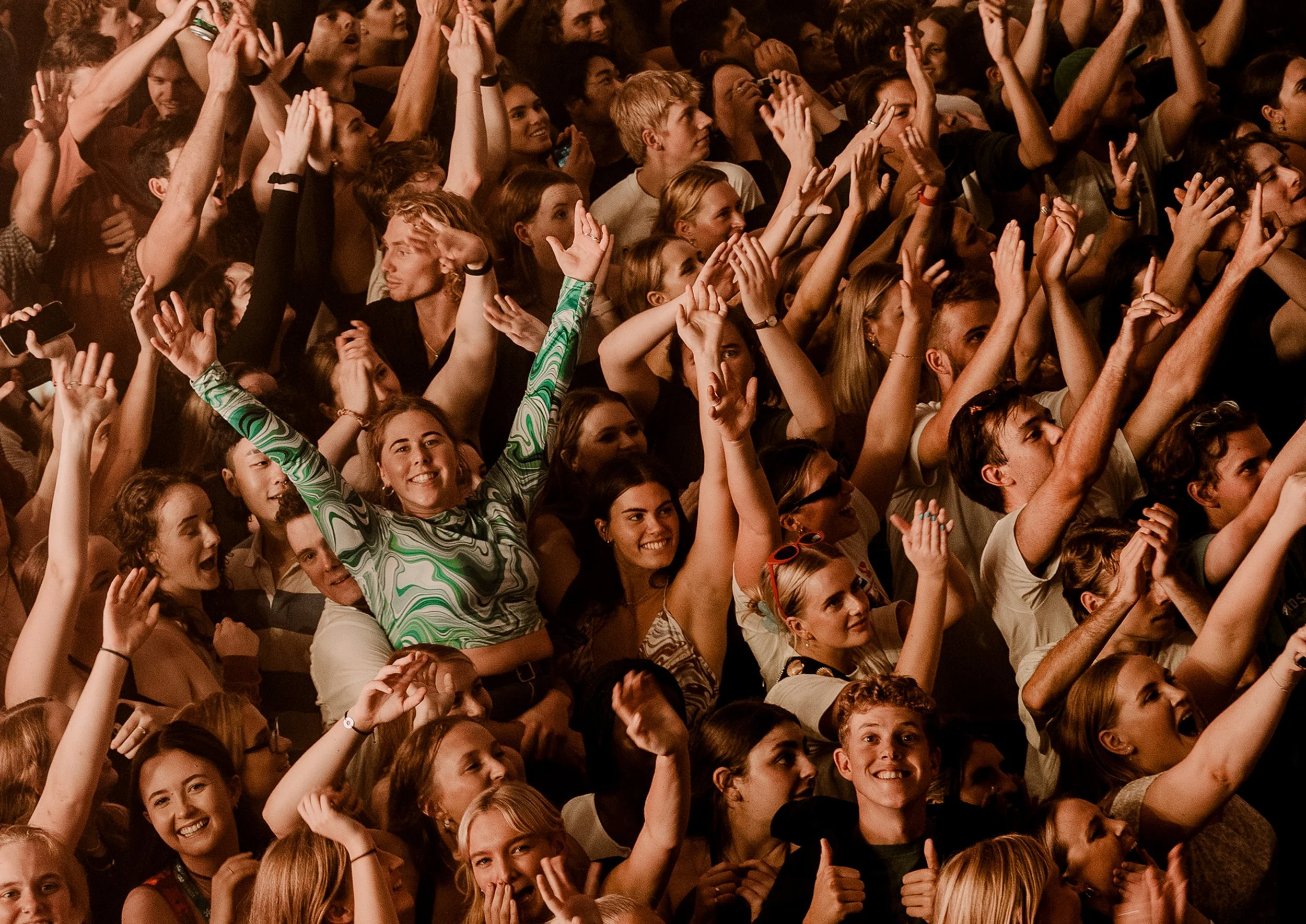 A large, enthusiastic crowd at a concert or event, raising their hands and smiling.