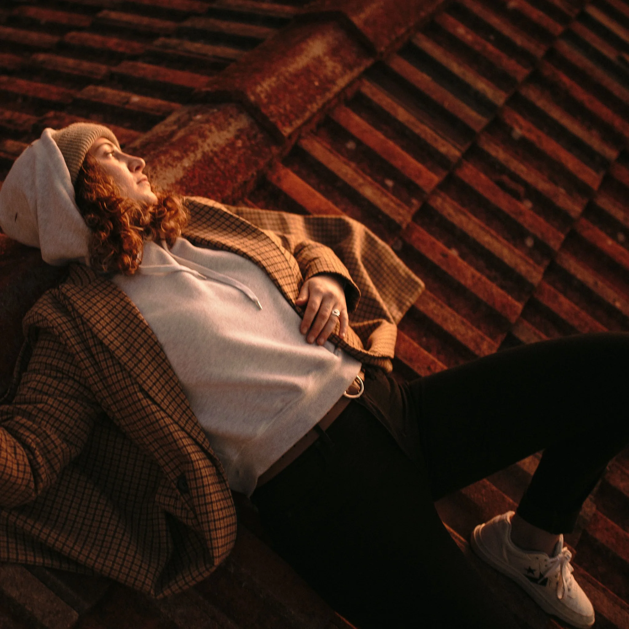 A person with curly hair wearing a khaki beanie and hoodie, lying on a bench with a checkered brown jacket, next to someone in white Converse sneakers, on a textured brick surface.
