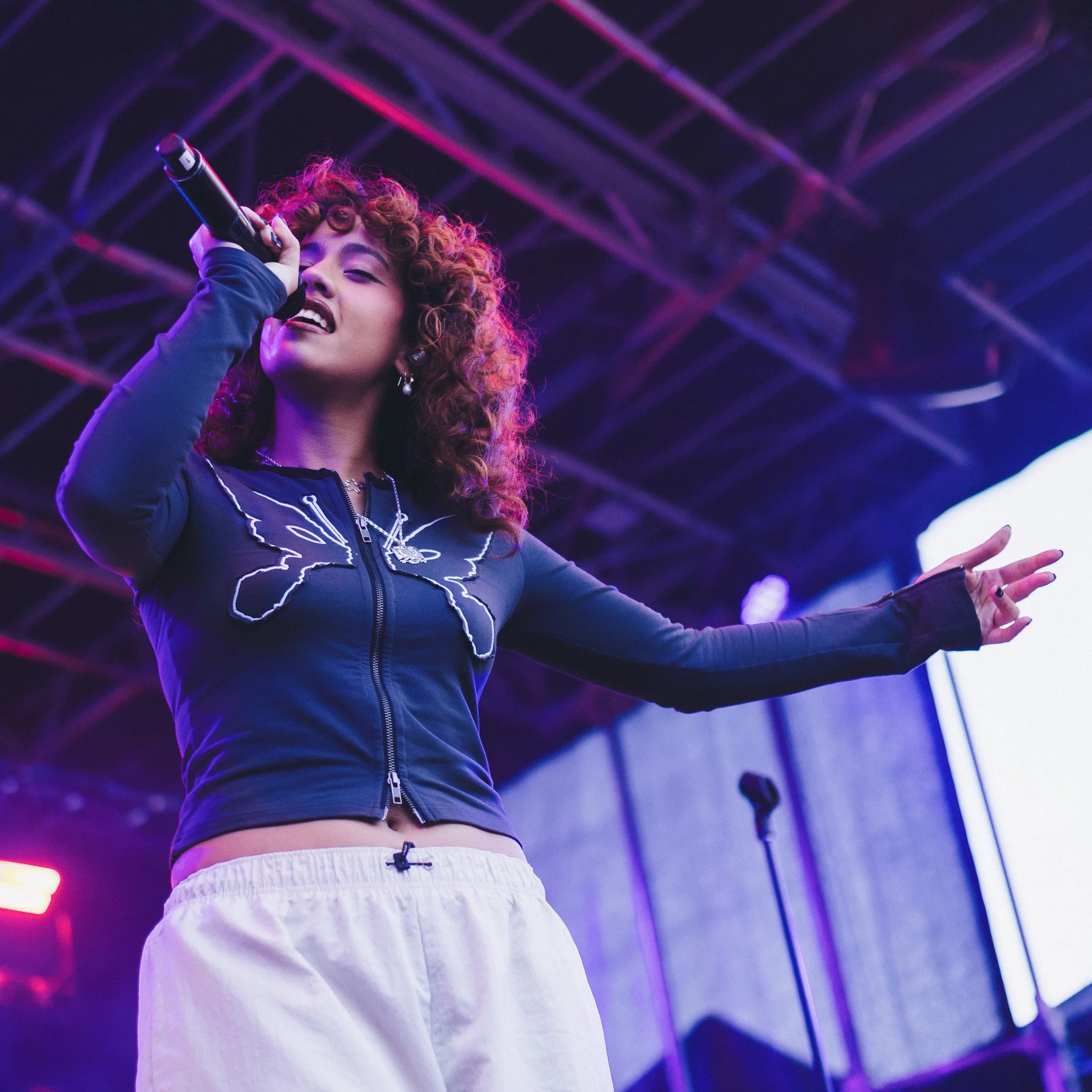 A young woman with curly hair joyfully singing or speaking into a microphone on a stage, illuminated by purple and blue stage lighting, with a metal truss structure in the background.