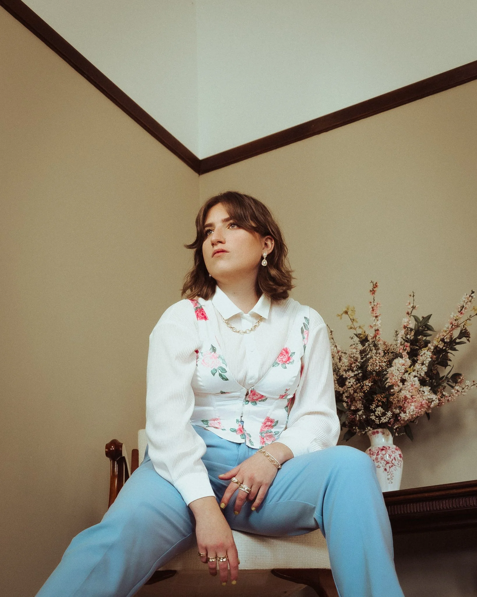 A woman with brown hair styled in waves, wearing a white blouse with floral embroidery, light blue pants, and decorative jewelry, is sitting on a chair in a room with beige walls, dark wooden crown molding, and a large bouquet of pink, white, and pur