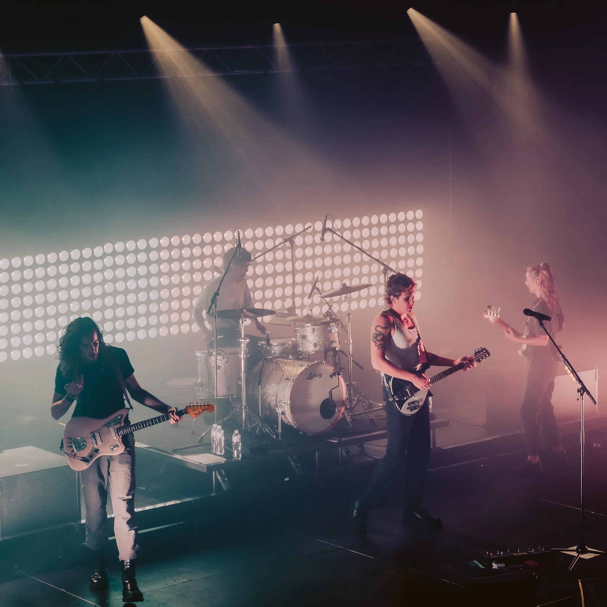 A live music concert scene featuring band members playing guitars and drums on stage with colorful lighting and a dotted light wall in the background.