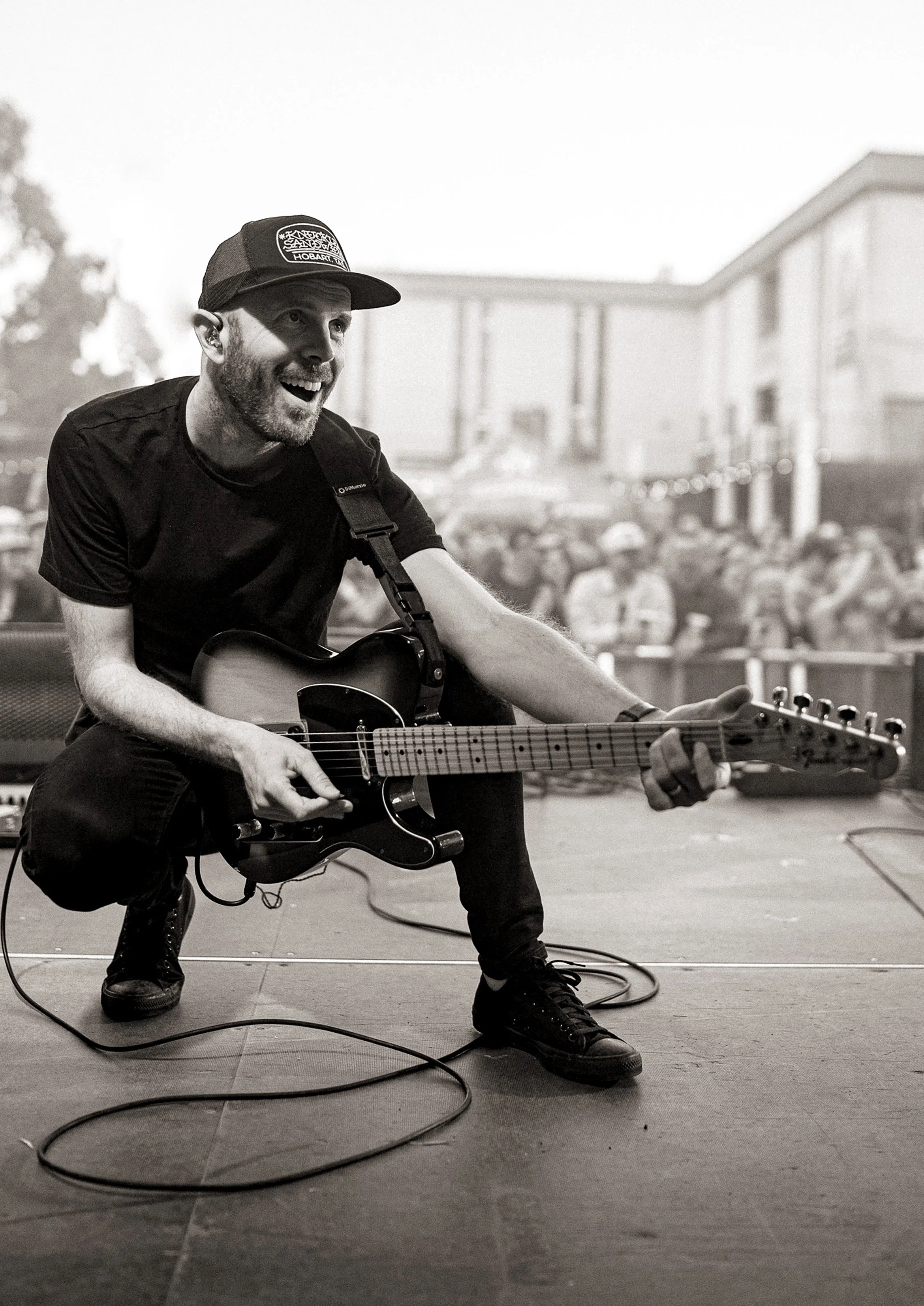 Black and white photo of a man playing an electric guitar on stage, smiling, wearing a black t-shirt, black shoes, and a baseball cap, with a crowd and a building in the background.