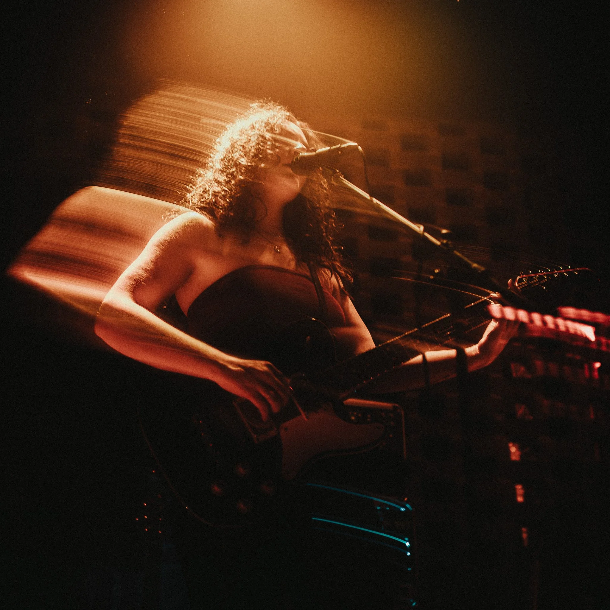 A woman with curly hair singing into a microphone during a live performance, with motion blur creating a dynamic light trail effect.