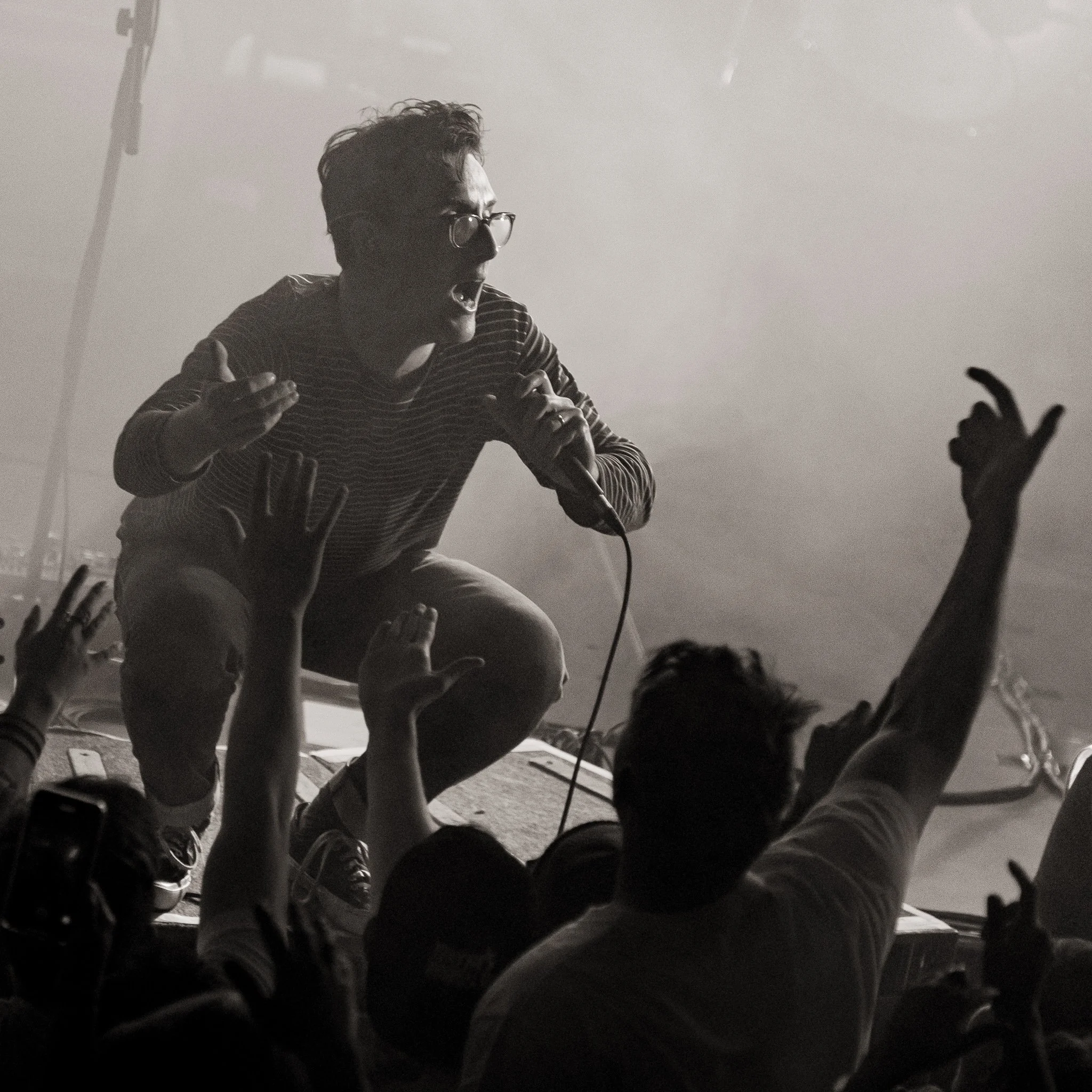 A person wearing glasses and a striped shirt is seen from below, with their mouth open and hand on their chest, surrounded by many hands reaching out towards them, in a dimly lit setting.