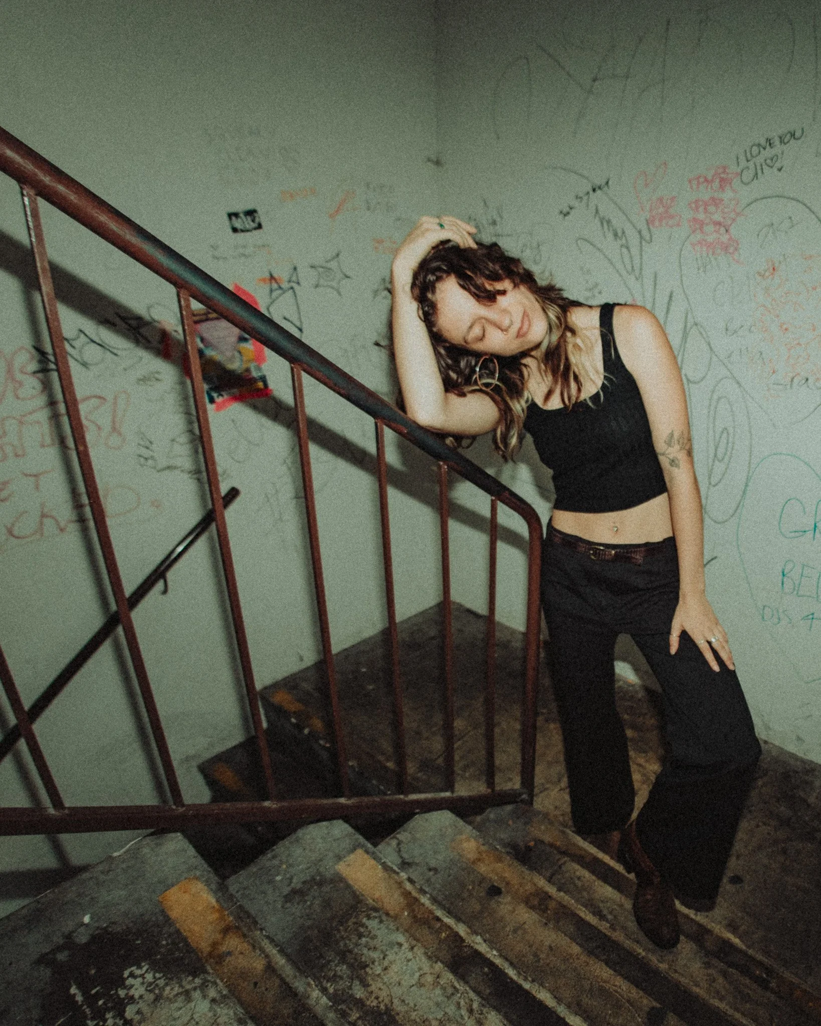A young woman with long, wavy hair standing on a staircase, leaning on a railing with her hand on her head. She is wearing a black crop top and black pants, with a belly button piercing and hoop earrings. The background features a wall with graffiti 