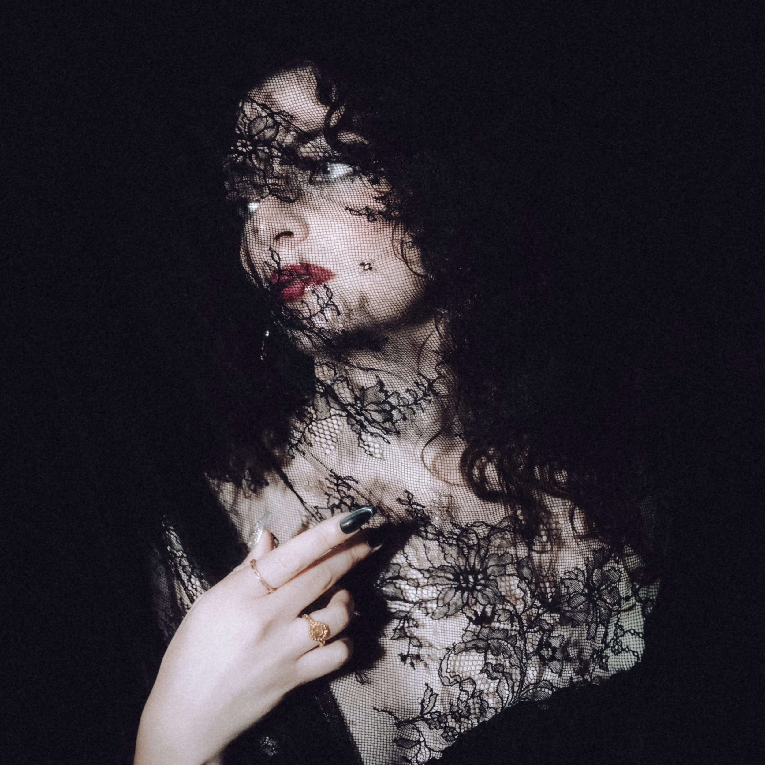 A woman with curly hair and makeup, wearing black lace fabric covering her face and neck, with her hand with black nail polish and rings visible.