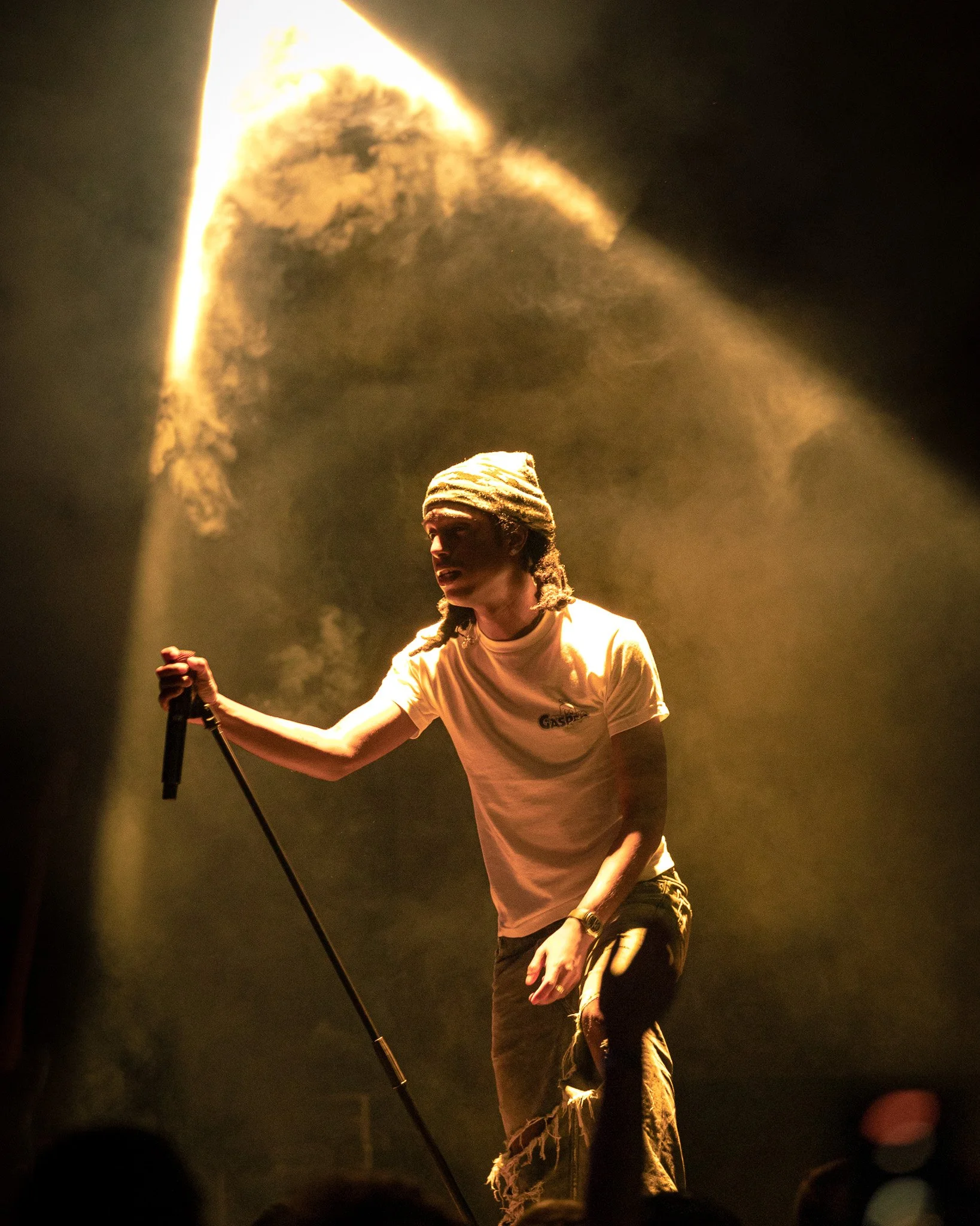 A performer on stage holding a microphone, wearing a light-colored T-shirt and a head covering, gesturing with one hand, illuminated by warm stage lighting, with a dark background.