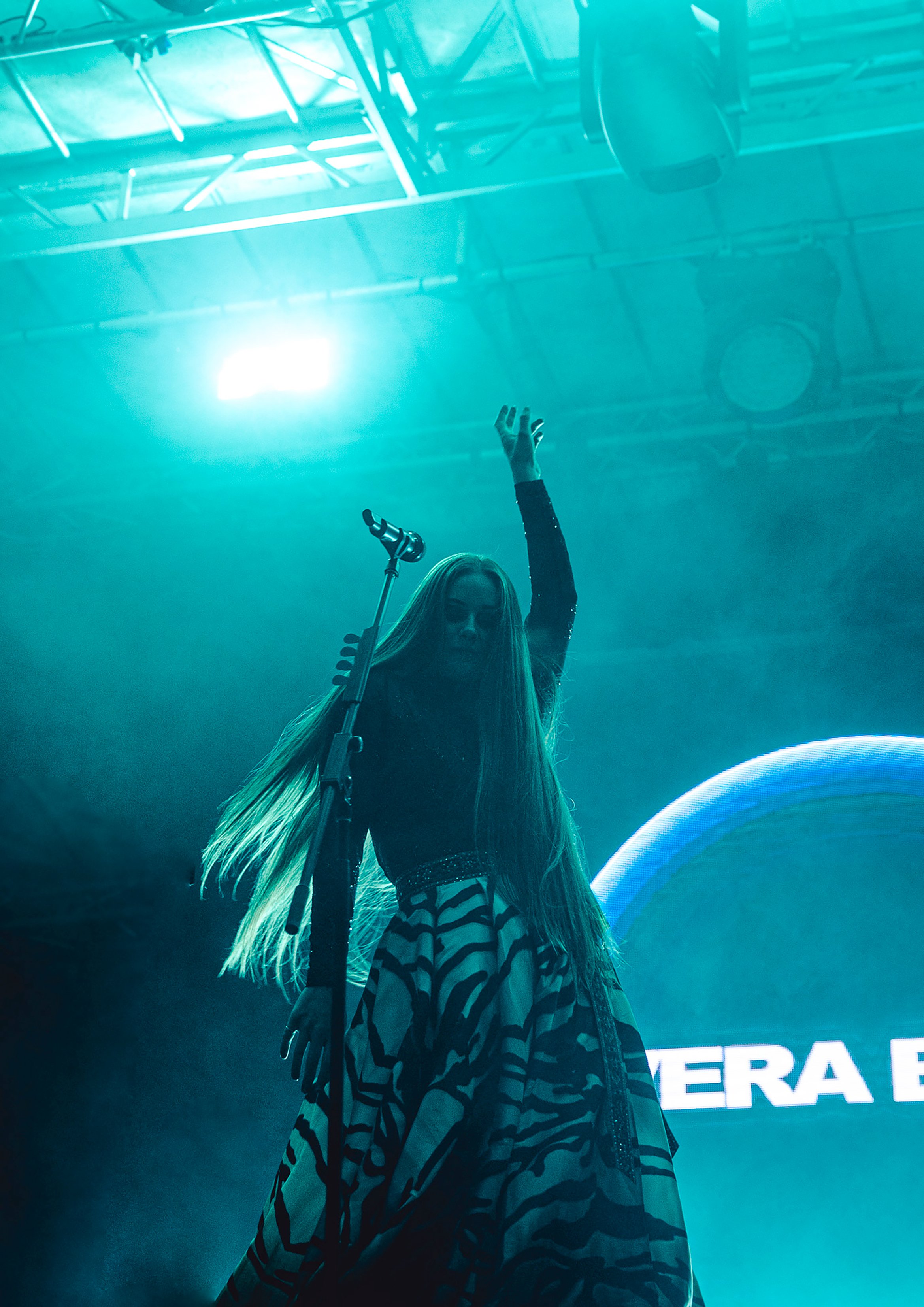 A woman standing on stage with a raised arm, microphone in front of her, and long blonde hair. The stage background is blue with part of a sign visible.