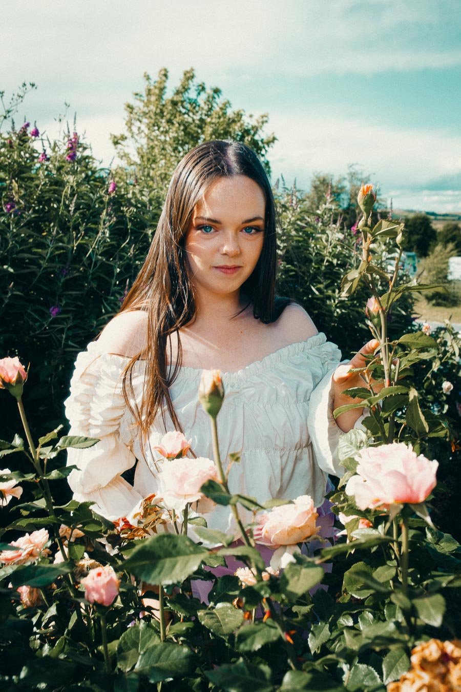 A young woman with long brown hair and blue eyes standing among pink roses in a garden on a sunny day, wearing an off-shoulder white dress.