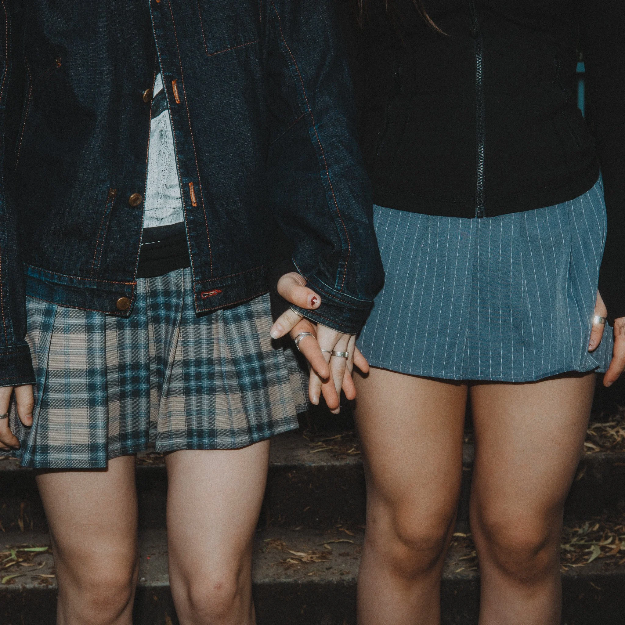 Four women standing side by side outdoors, showing their legs from knees to feet, wearing denim skirts and shorts, with some making hand gestures.