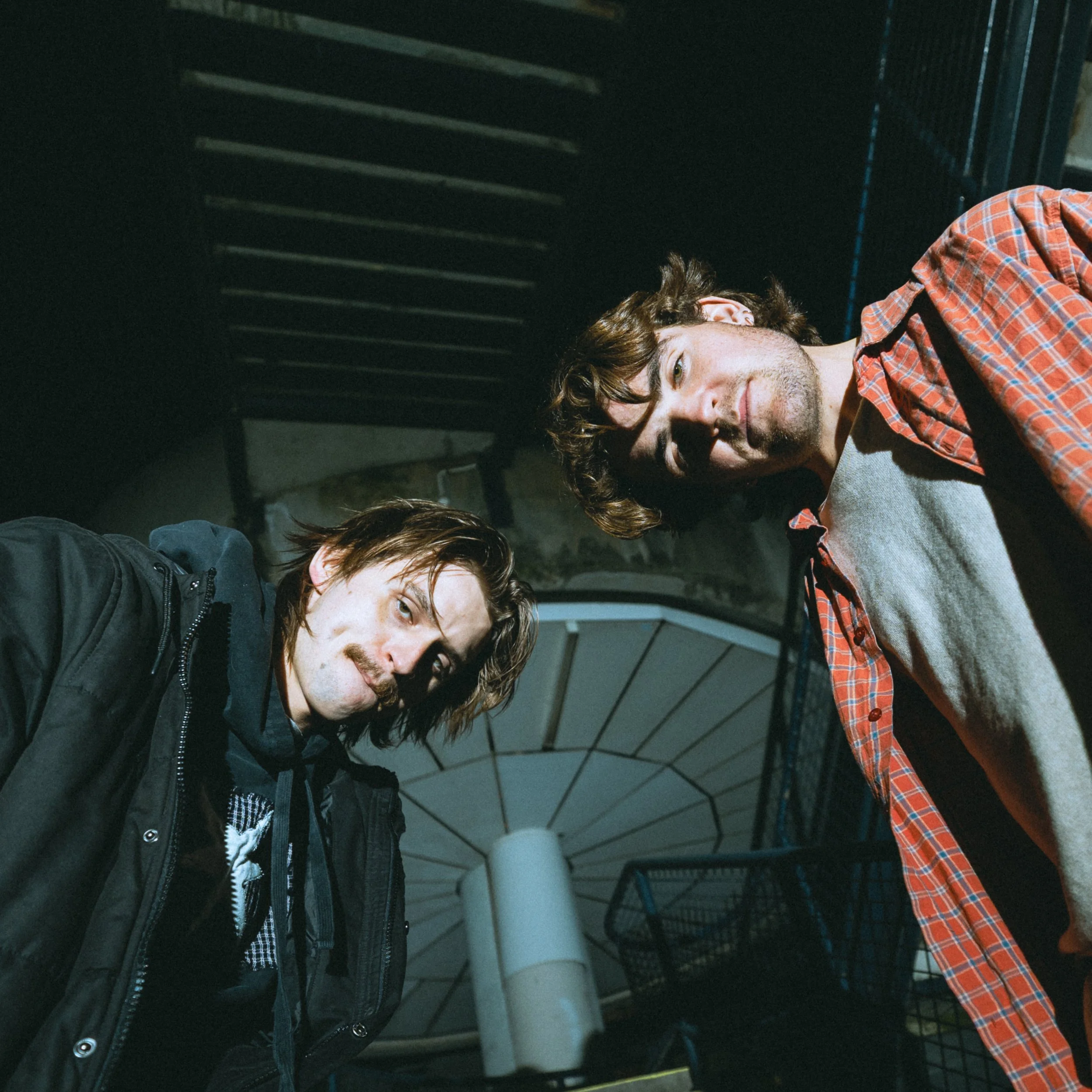 Three young men are sitting on a metal staircase, looking at the camera. They have casual hairstyles and are wearing casual clothing, including jackets and a plaid shirt. The staircase is indoors and painted black.