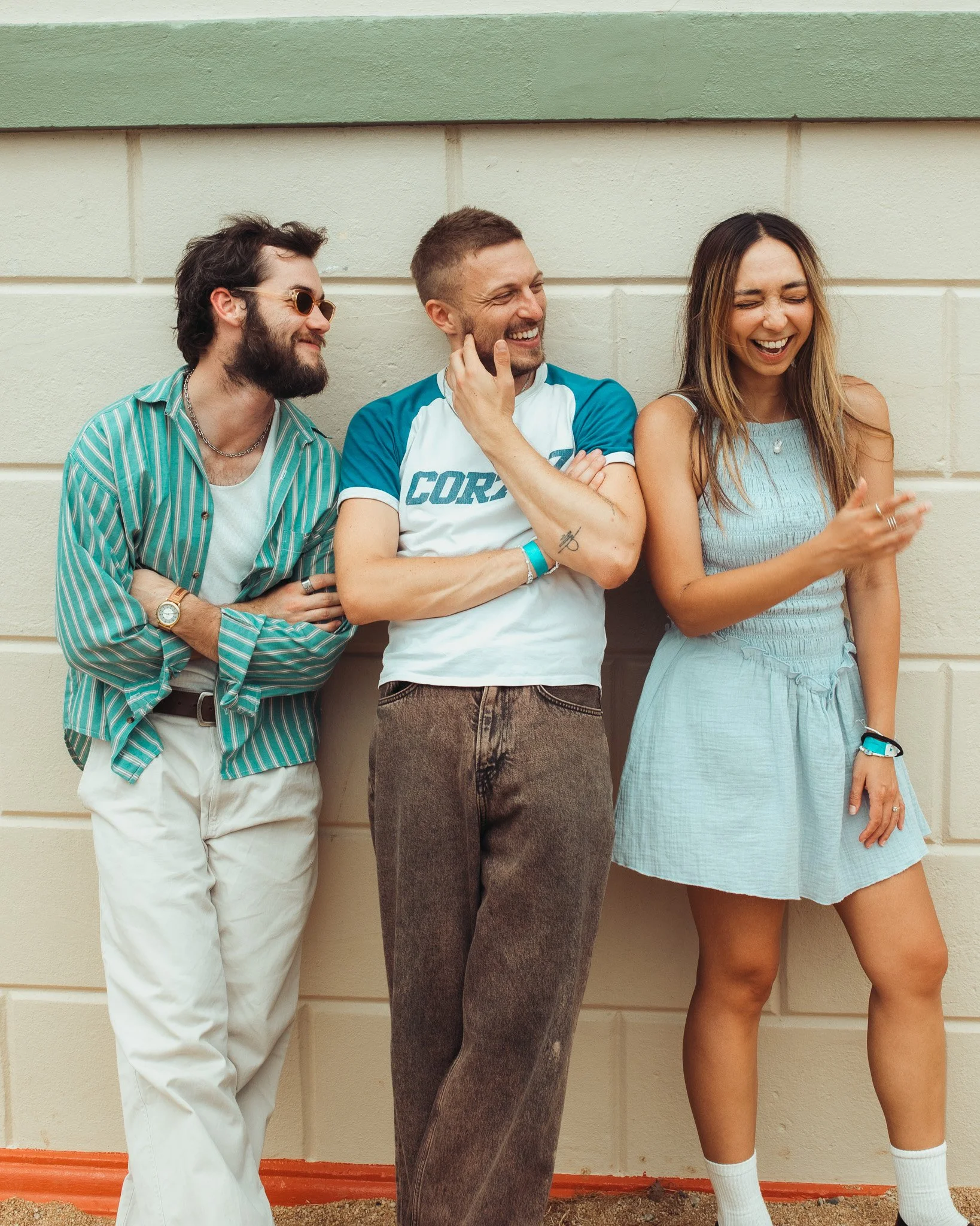 Three friends in casual clothes laughing and smiling together against a pastel-colored wall.