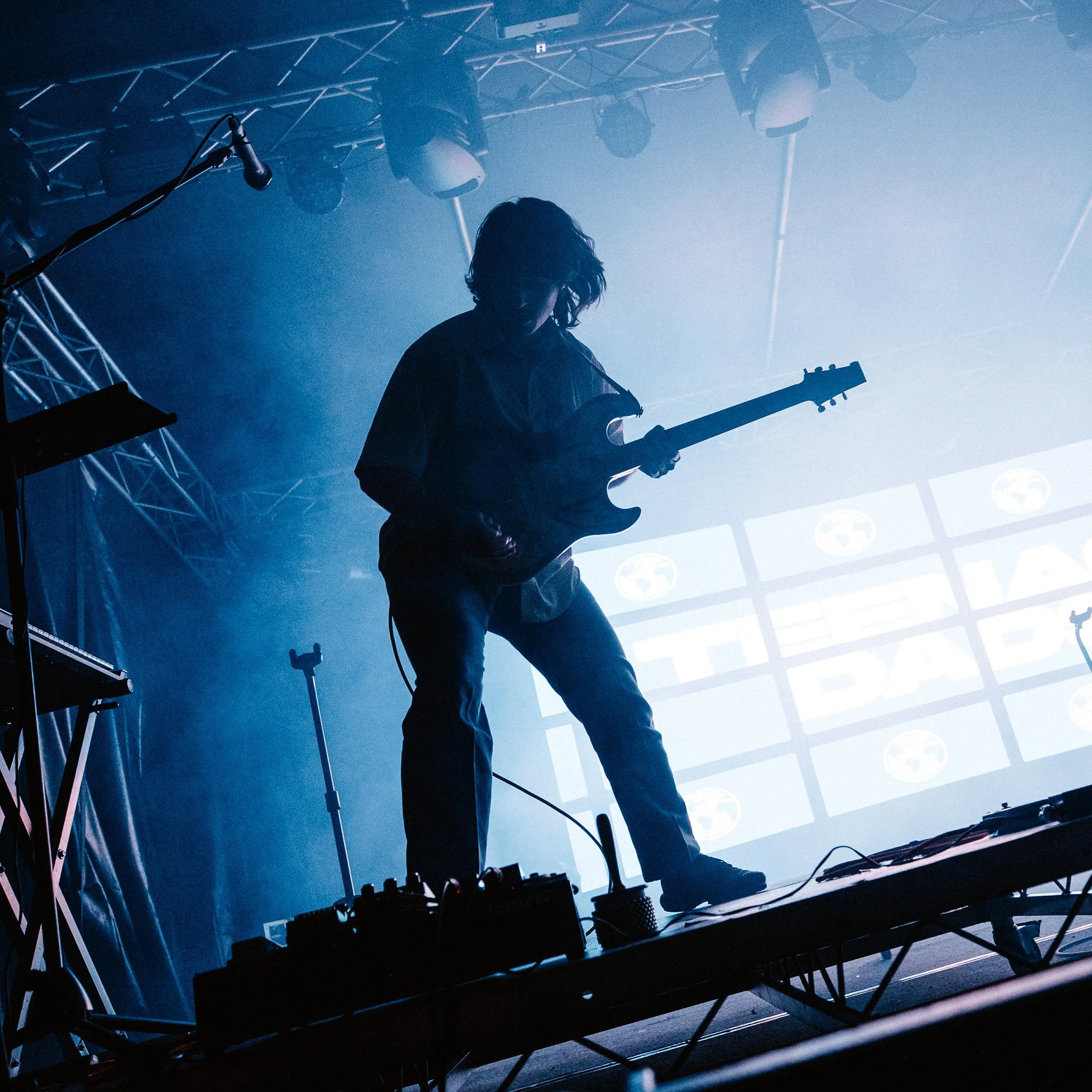 A musician with long hair playing an electric guitar on stage, silhouetted against stage lights and a foggy atmosphere, with equipment and instruments visible in the background.