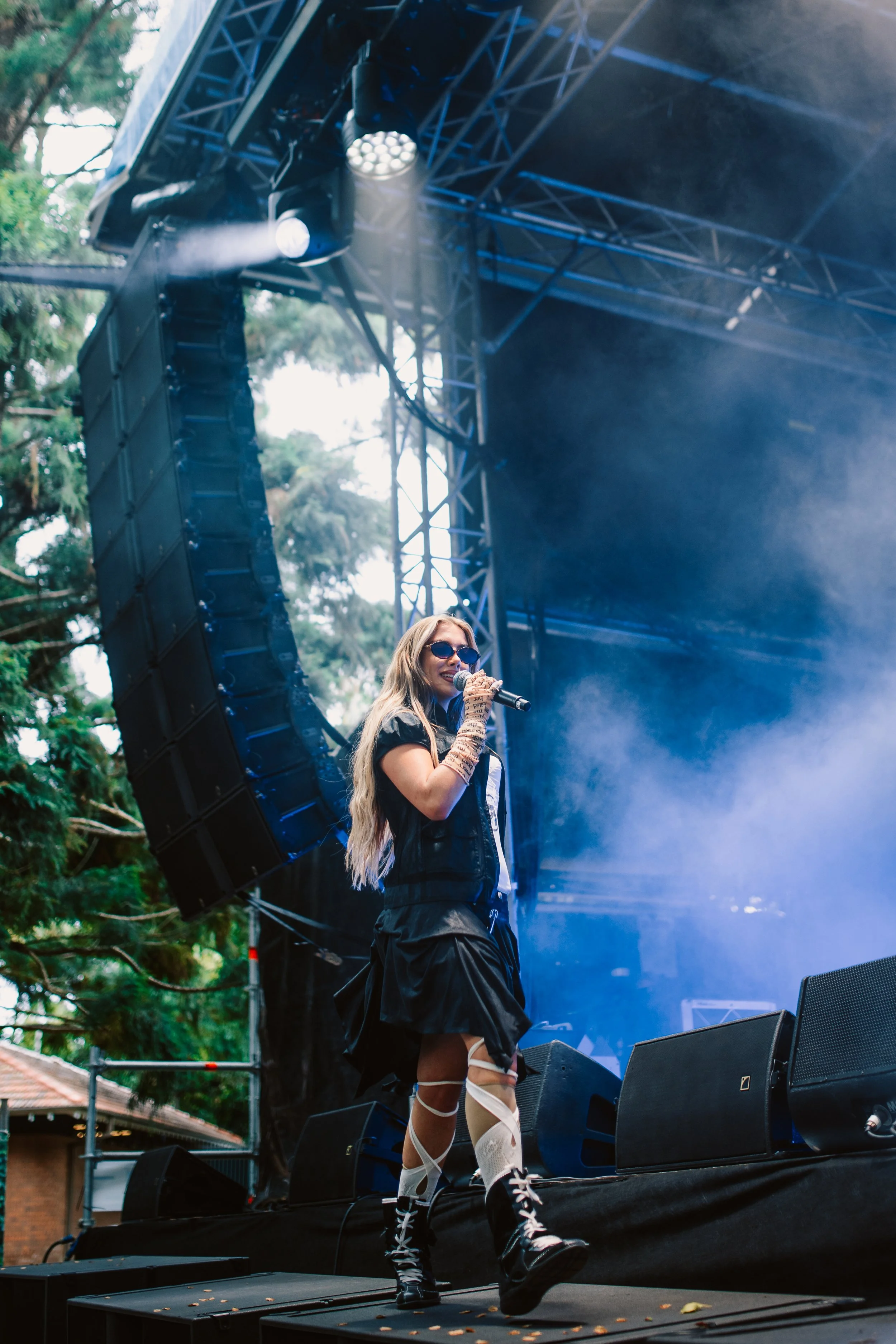 A female performer with long blonde hair, sunglasses, and patterned thigh-high stockings singing into a microphone on an outdoor stage during daylight, with large speakers and stage equipment around her.