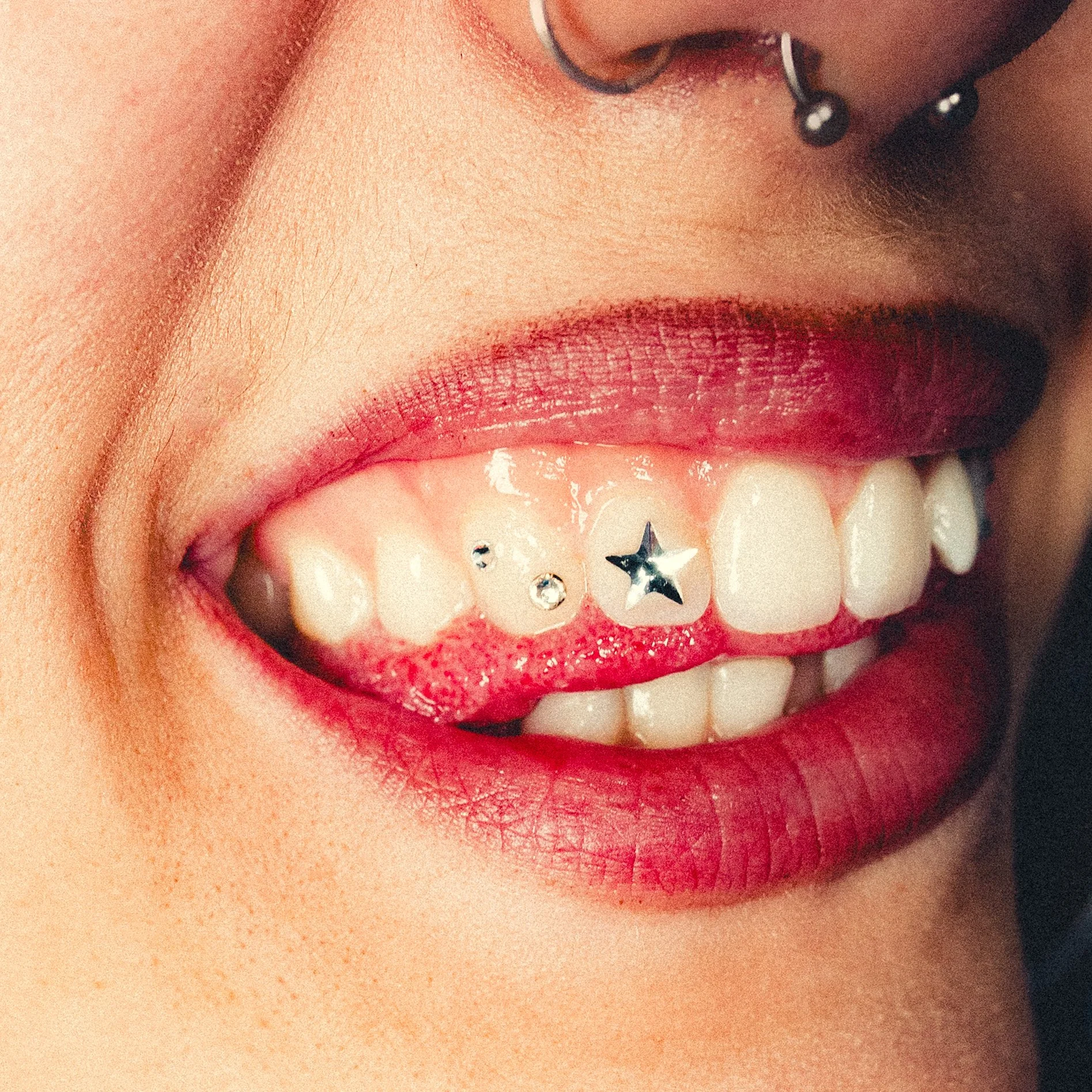 Close-up of a person's mouth with lipstick and a nose piercing, showing teeth and some jewelry on the lips.