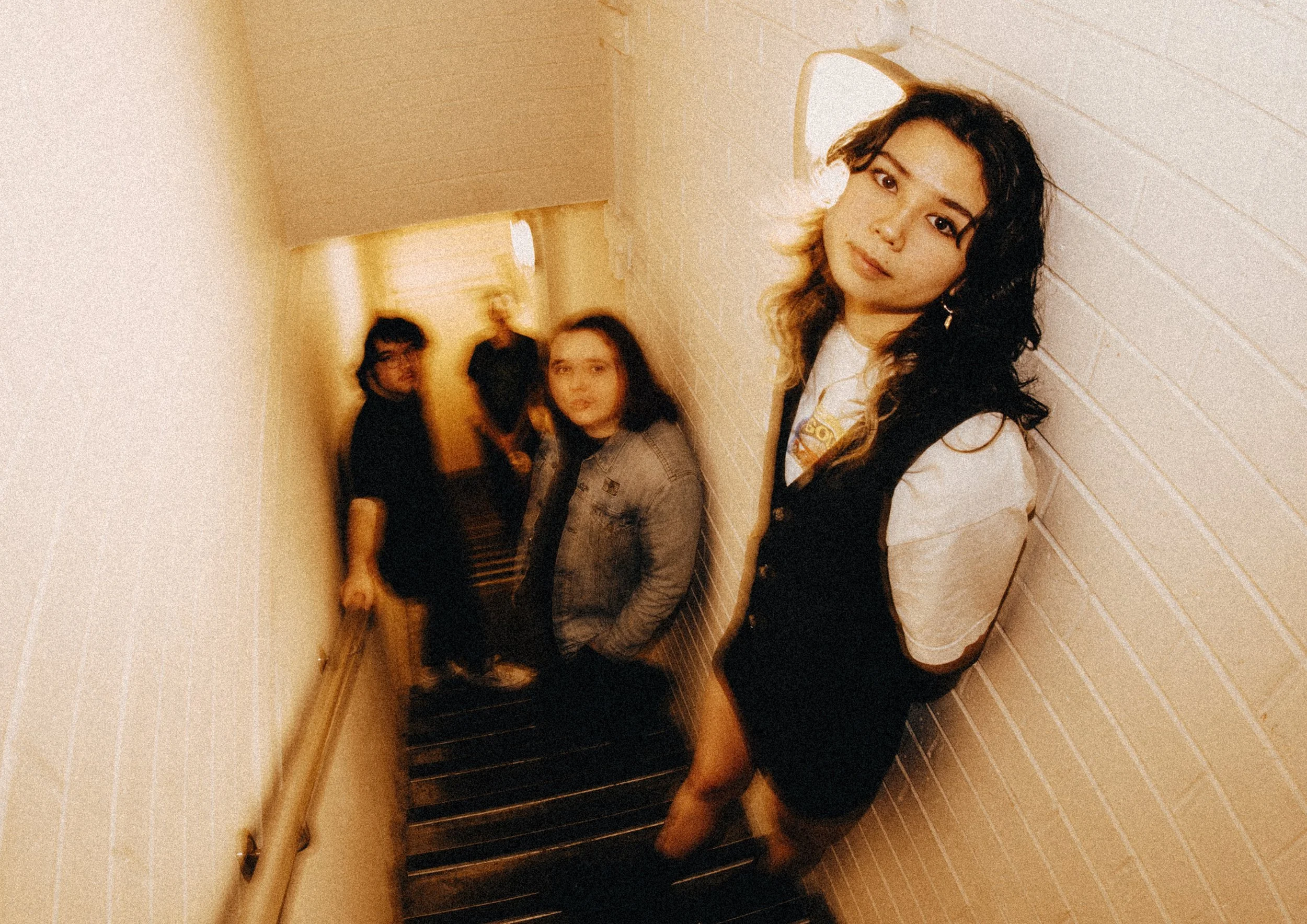 A group of four young women standing in a hallway, with one woman in the foreground leaning against the wall and the others further back, all looking at the camera.