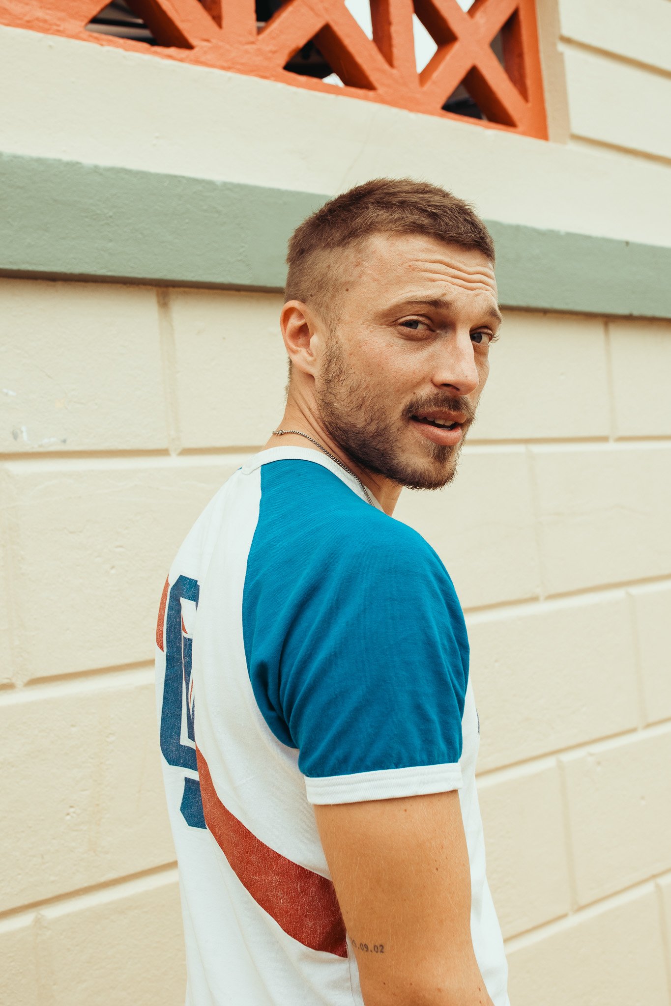 A man with short brown hair, beard, and light skin tone, wearing a blue and white shirt, standing in front of a beige wall with a red decorative brick pattern near the top, looking over his shoulder.