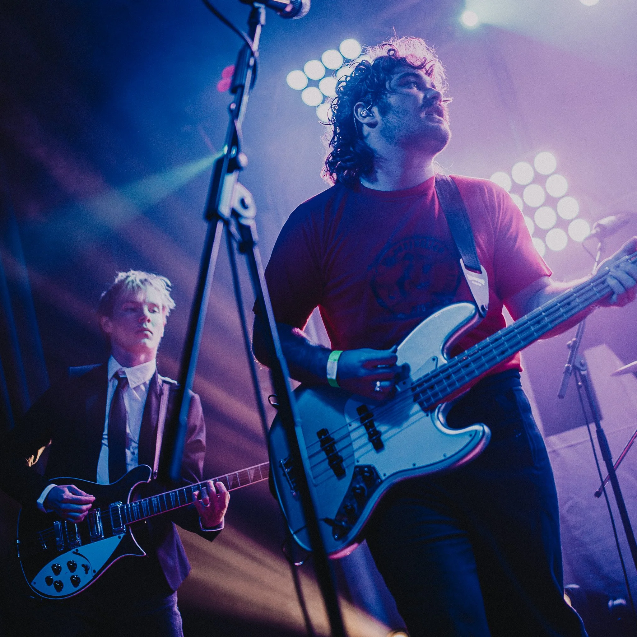 A musician playing an electric bass guitar on stage under bright, colorful lights, with another band member in the background.
