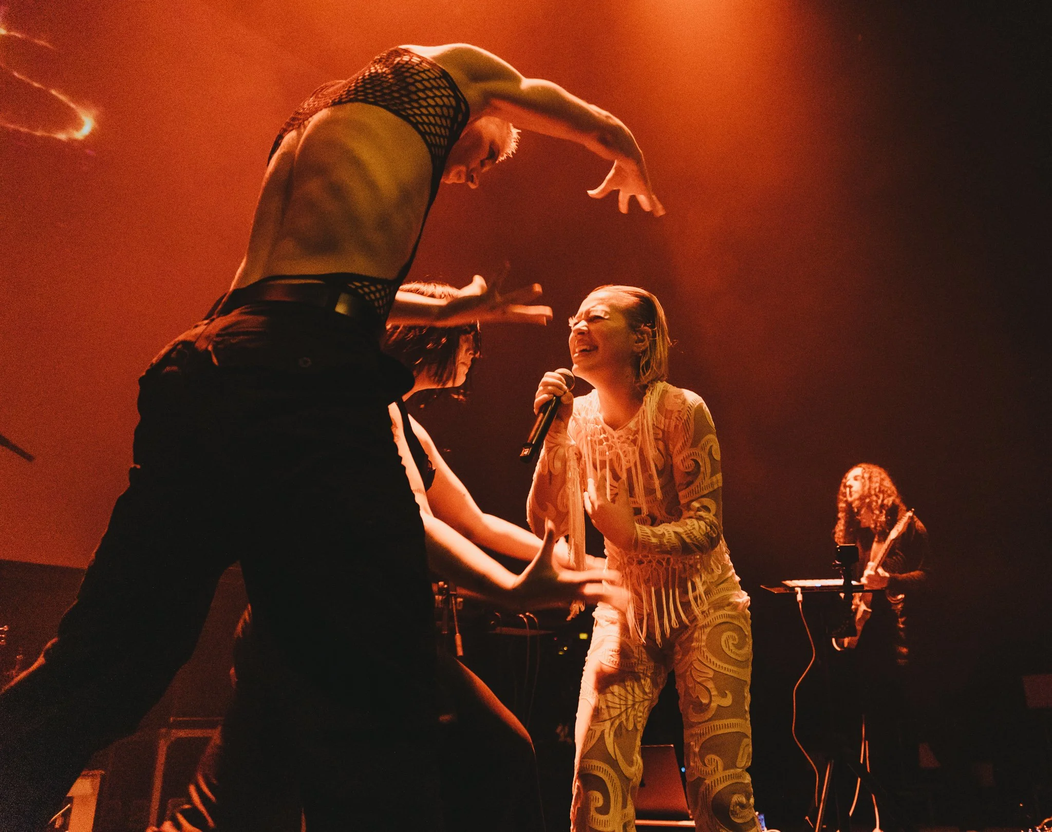 A group of people dancing and singing on stage, with a woman in a yellow dress holding a microphone, illuminated by warm stage lighting.