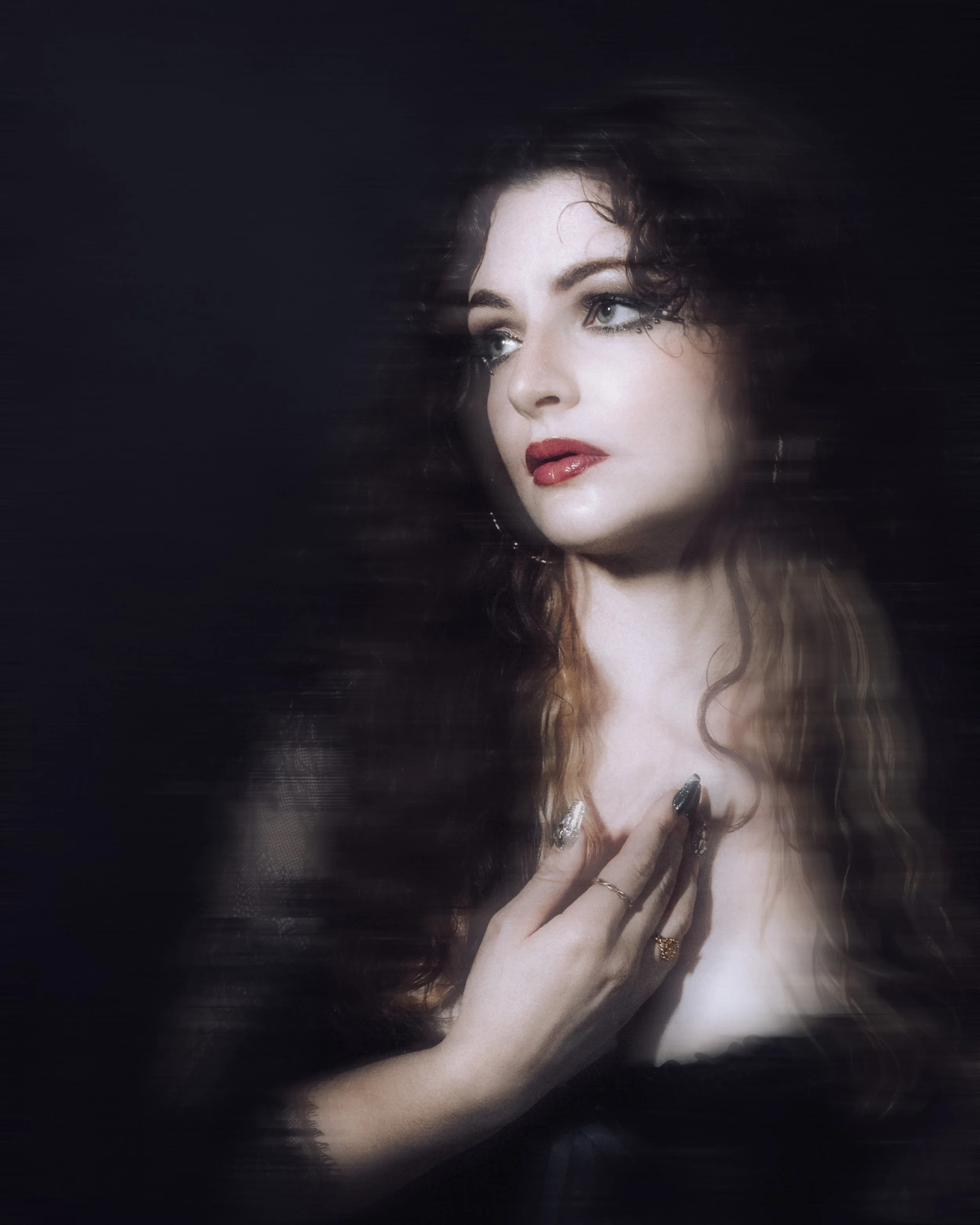A woman with dark wavy hair and makeup featuring red lipstick and eye embellishments, touching her neck with her left hand, adorned with rings, against a dark background.