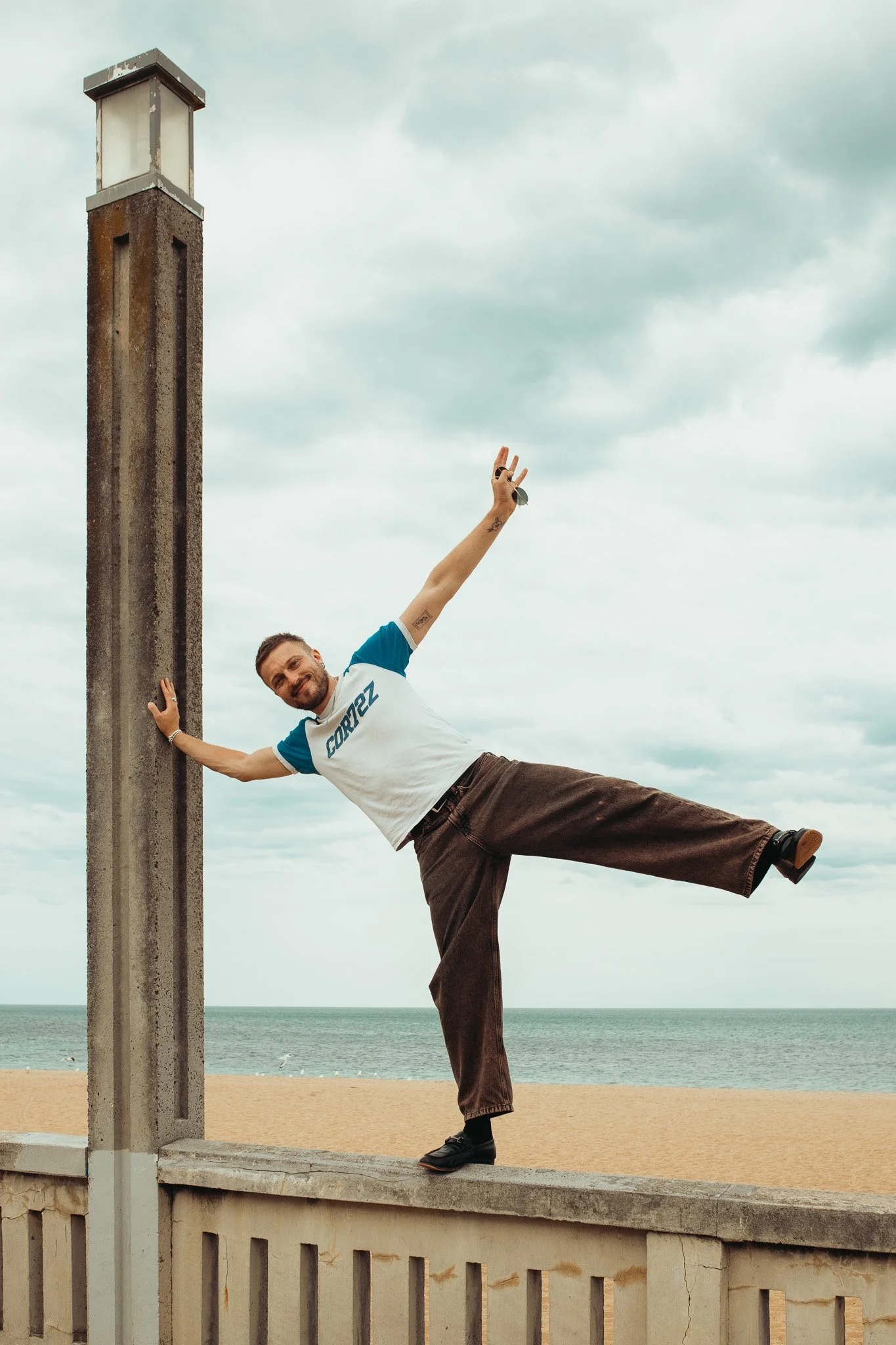 A man standing on a concrete wall by the beach, balancing on one leg with his other leg extended sideways, and holding onto a post for support. He is smiling and reaching his arm upward, holding sunglasses, while wearing a white and blue shirt with t